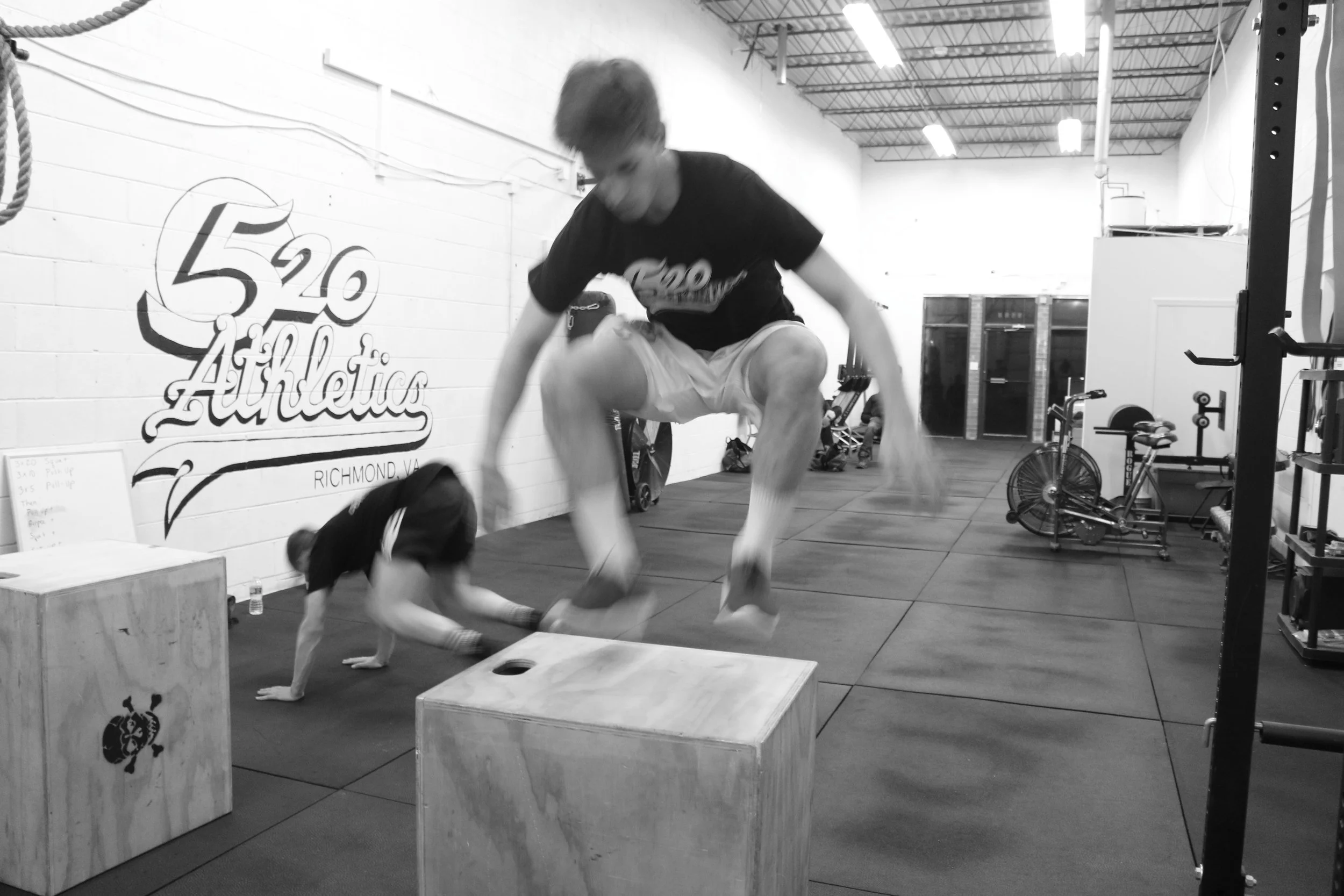 A person in mid-air jumping over a wooden plyometric box during a workout at a gym. The gym has equipment, bicycles, and a painted wall with the text "5 20 Athletics RICHMOND VA." The photo is in black and white.