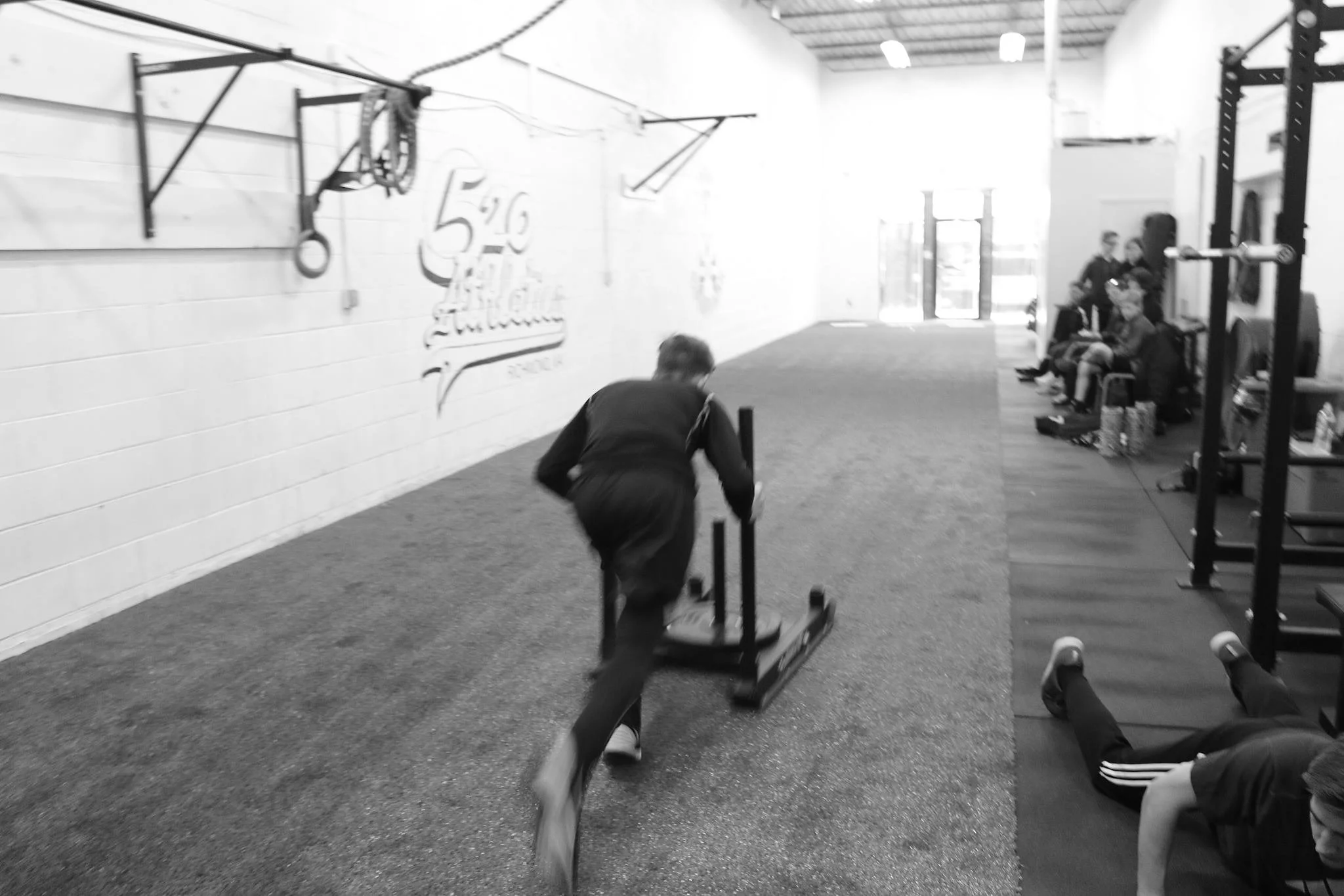 A person pushing a sled at the gym, with others seated on benches and lying on exercise mats nearby.