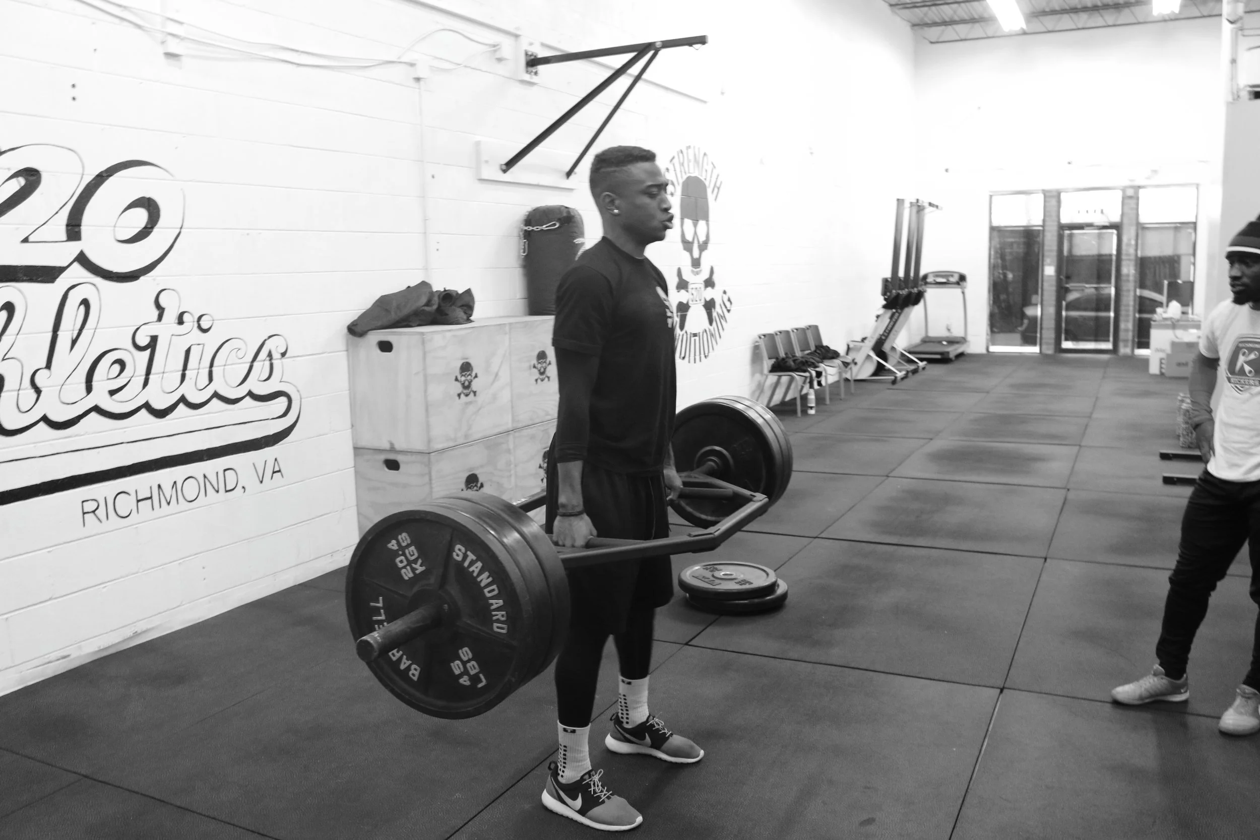Young man performing a deadlift with a barbell and weight plates in a gym. He is wearing a black t-shirt, black shorts, and sneakers. There is a trainer or coach observing on the right. The gym has a branded wall with the words "Richmond VA" and fitness-themed decorations.