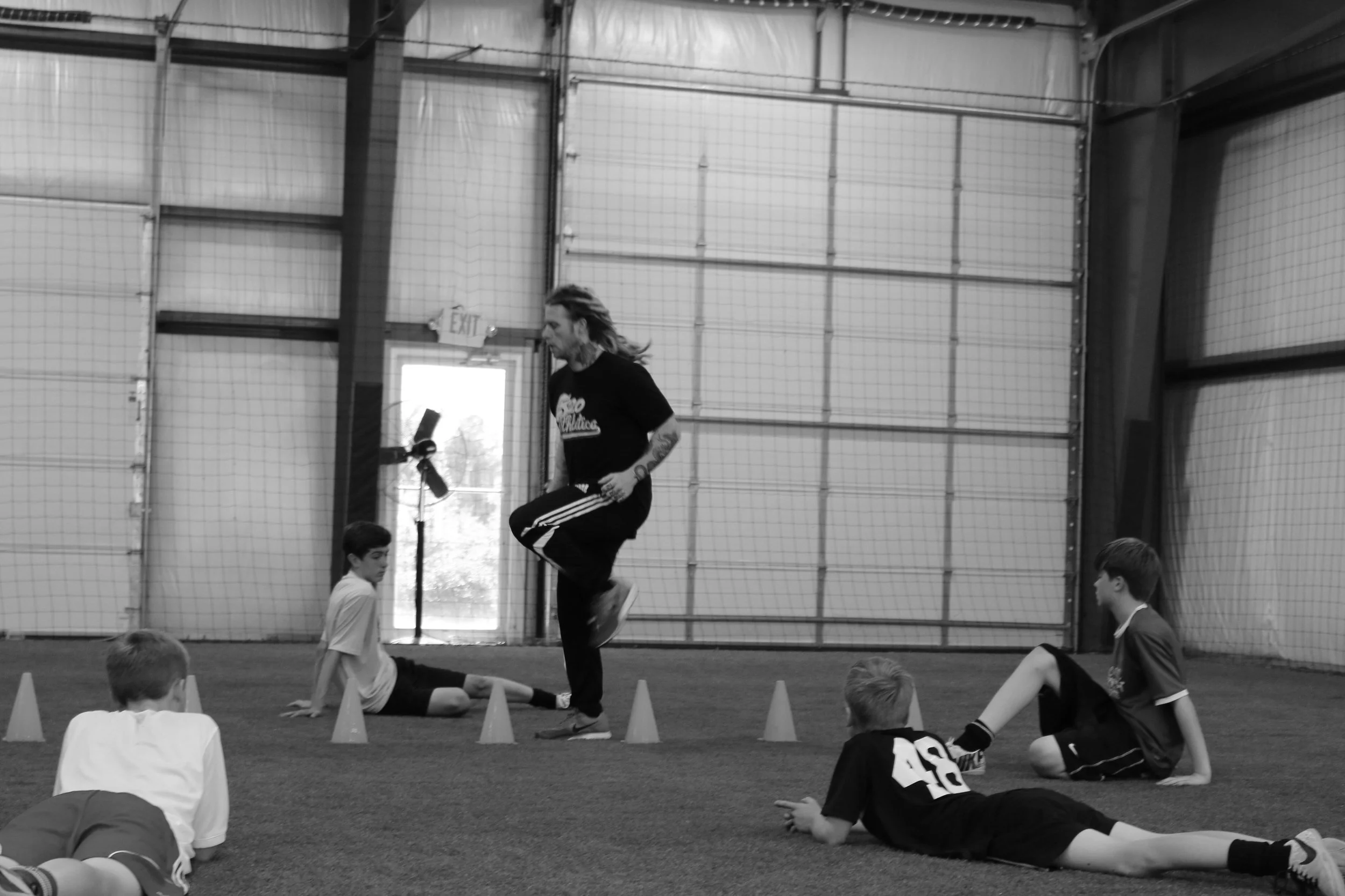A coach demonstrating a jump to a group of young boys during a training session inside an indoor sports facility.