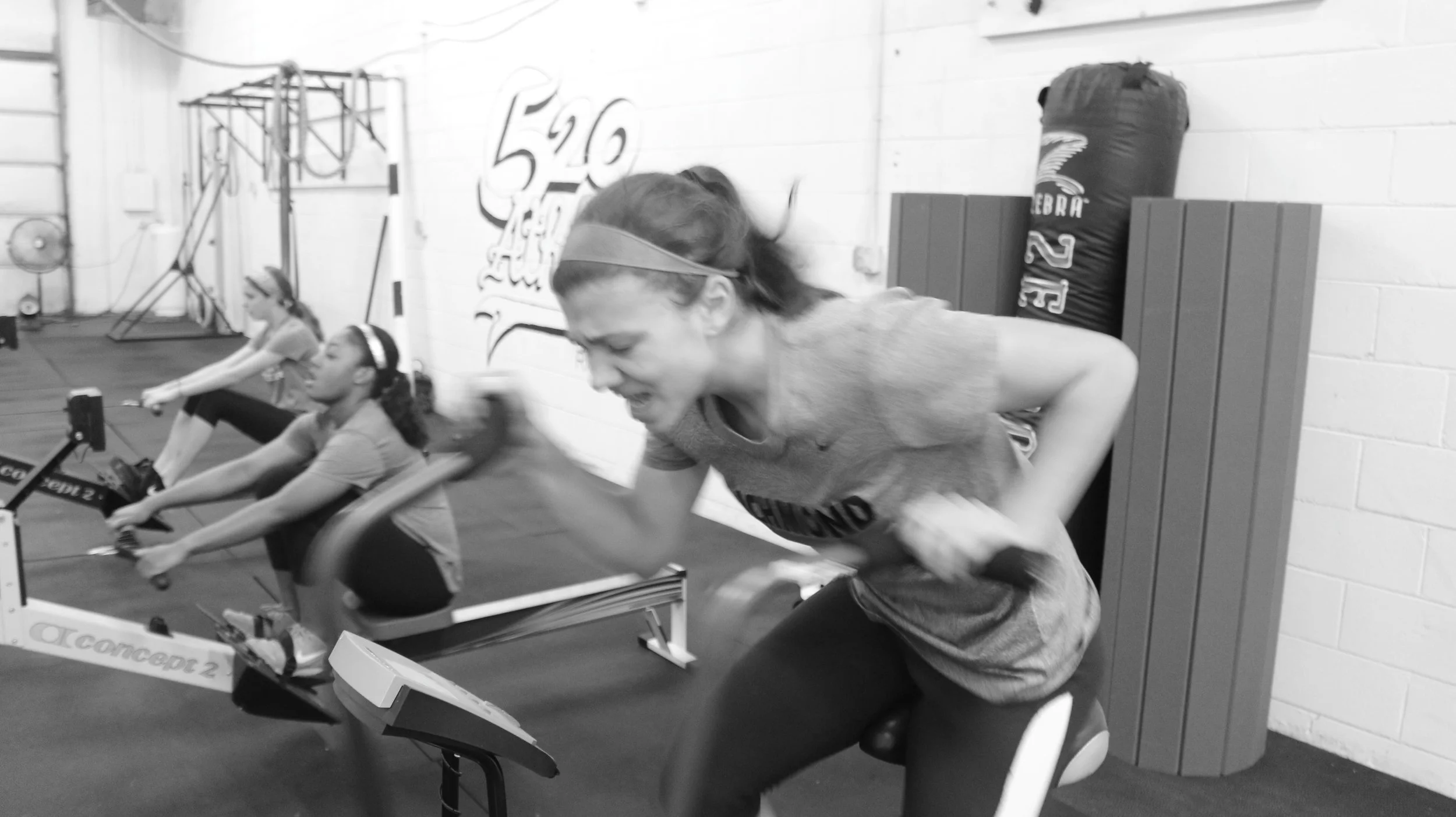 Three women working out on rowing machines in a gym, one in the foreground with a headband, appears to be tired or exerting effort.