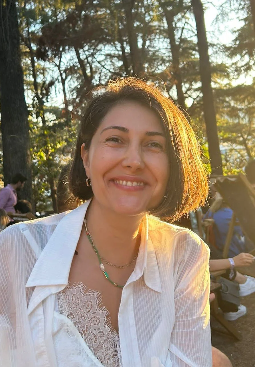 A woman with short brown hair smiling outdoors in front of trees during sunset, wearing a white blouse and layered necklaces.
