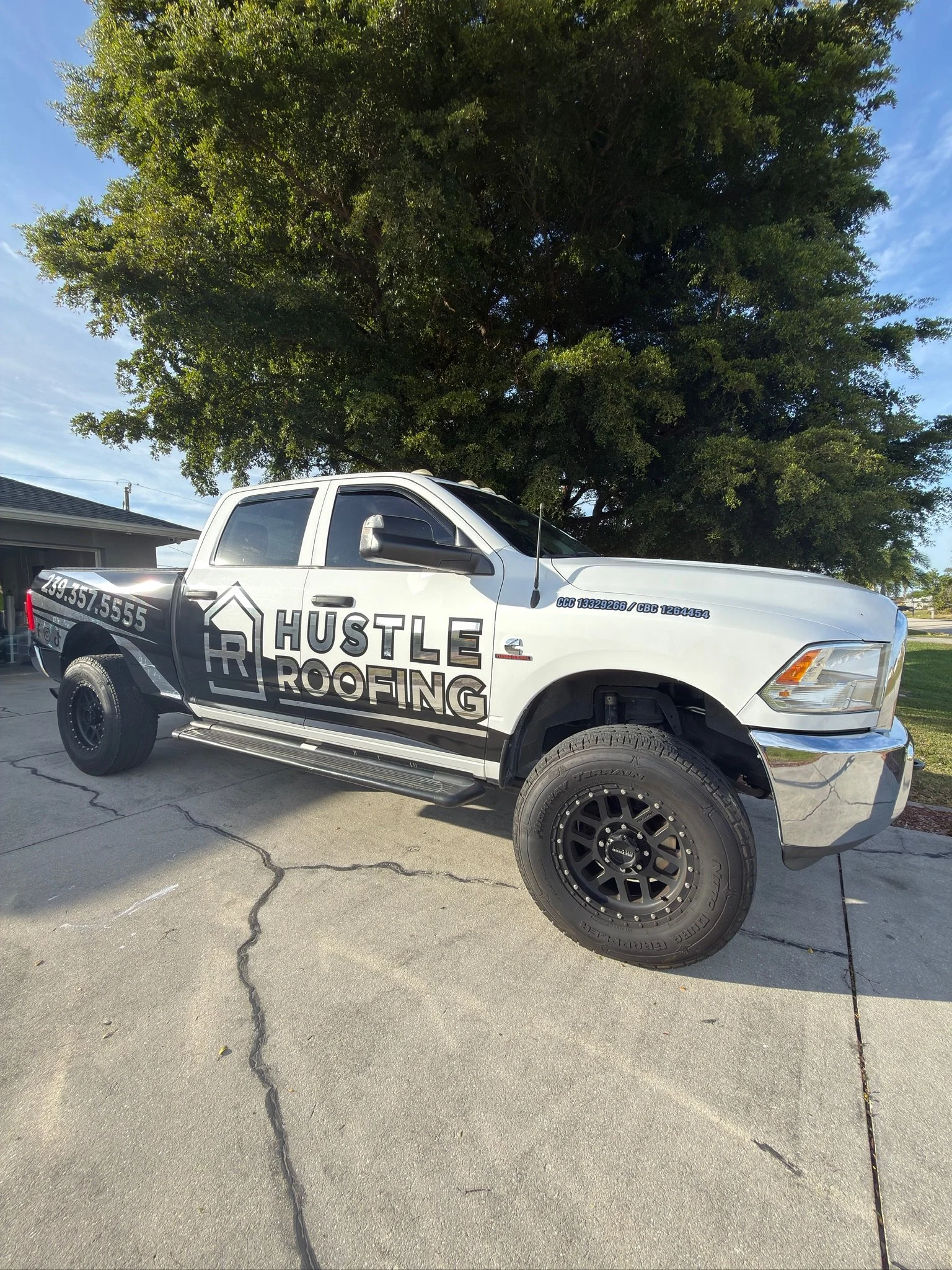 White pickup truck with black and silver Hustle Roofing logo and website on the side, parked on a concrete driveway in front of a large green tree, under a blue sky.