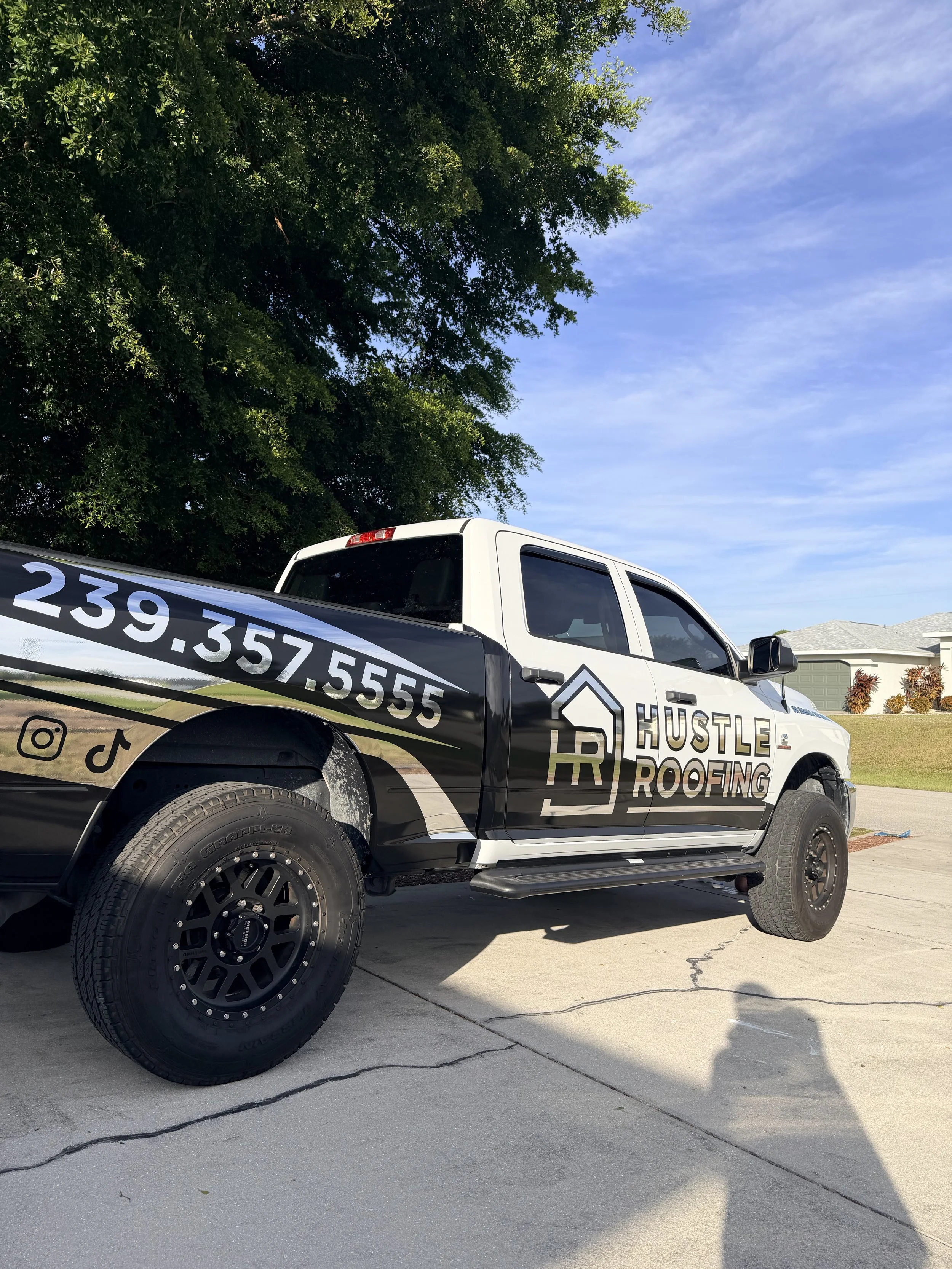 A pickup truck parked on a concrete driveway, decorated with black, white, silver graphics advertising 'Hustle Roofing' with contact information. The truck has black wheels and is parked next to a grassy area with trees and houses in the background.