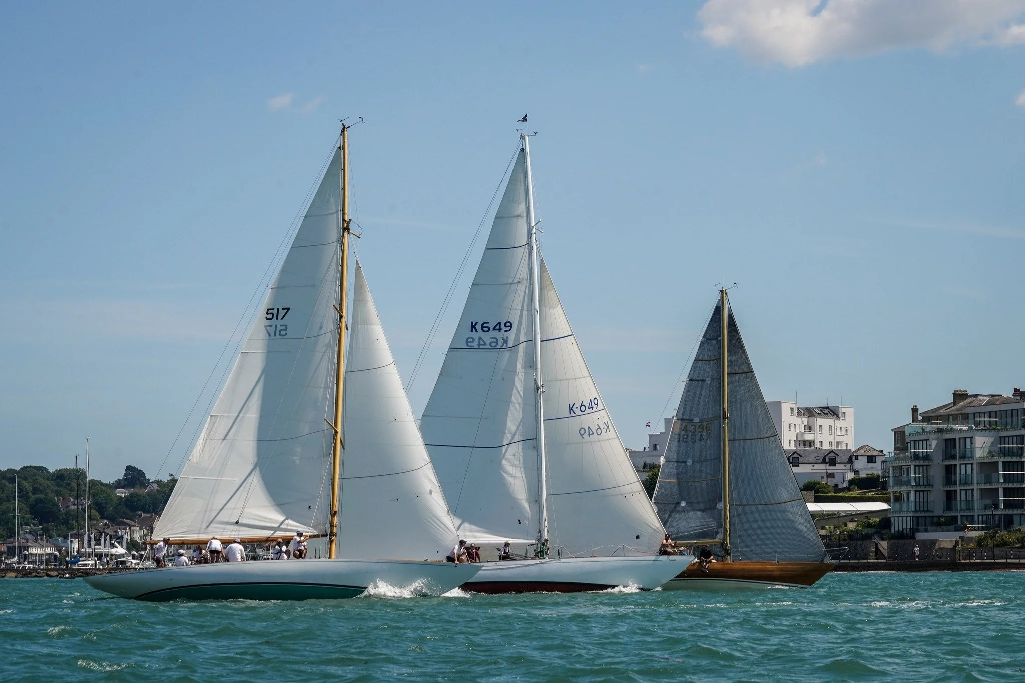 Three sailboats with white and dark sails racing on the water near a shoreline with modern buildings under a partly cloudy sky.