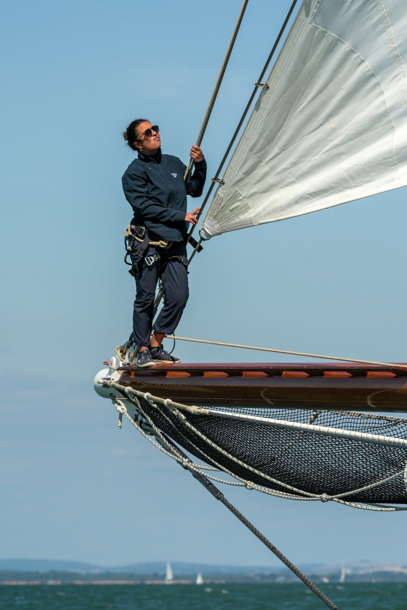 A woman standing on the bow of a sailing boat, holding the mast with her right hand, while sailing on the water under a clear sky.