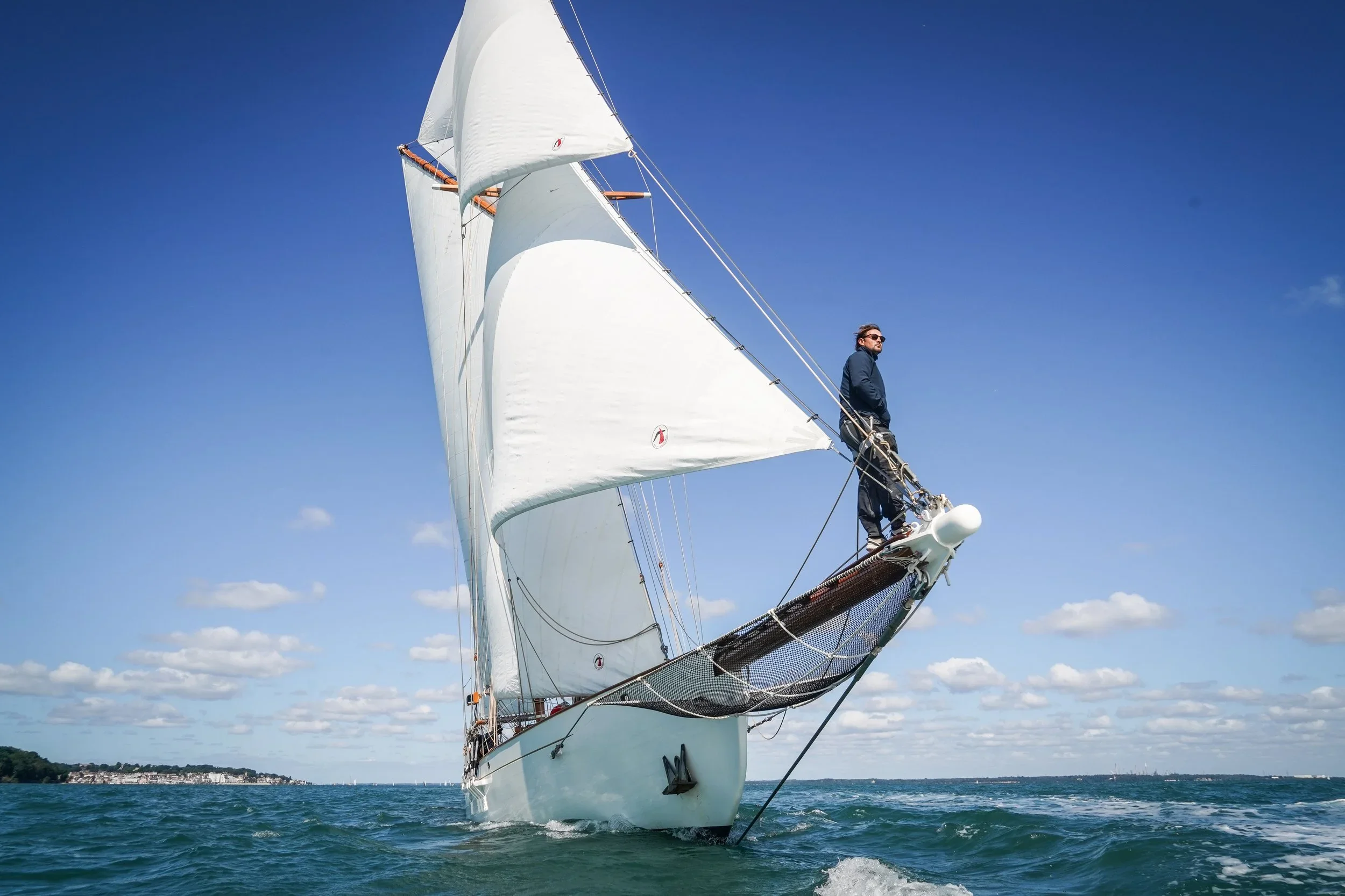 A large white sailing yacht with a man standing on the bow, sailing on the ocean under a clear blue sky with scattered clouds.