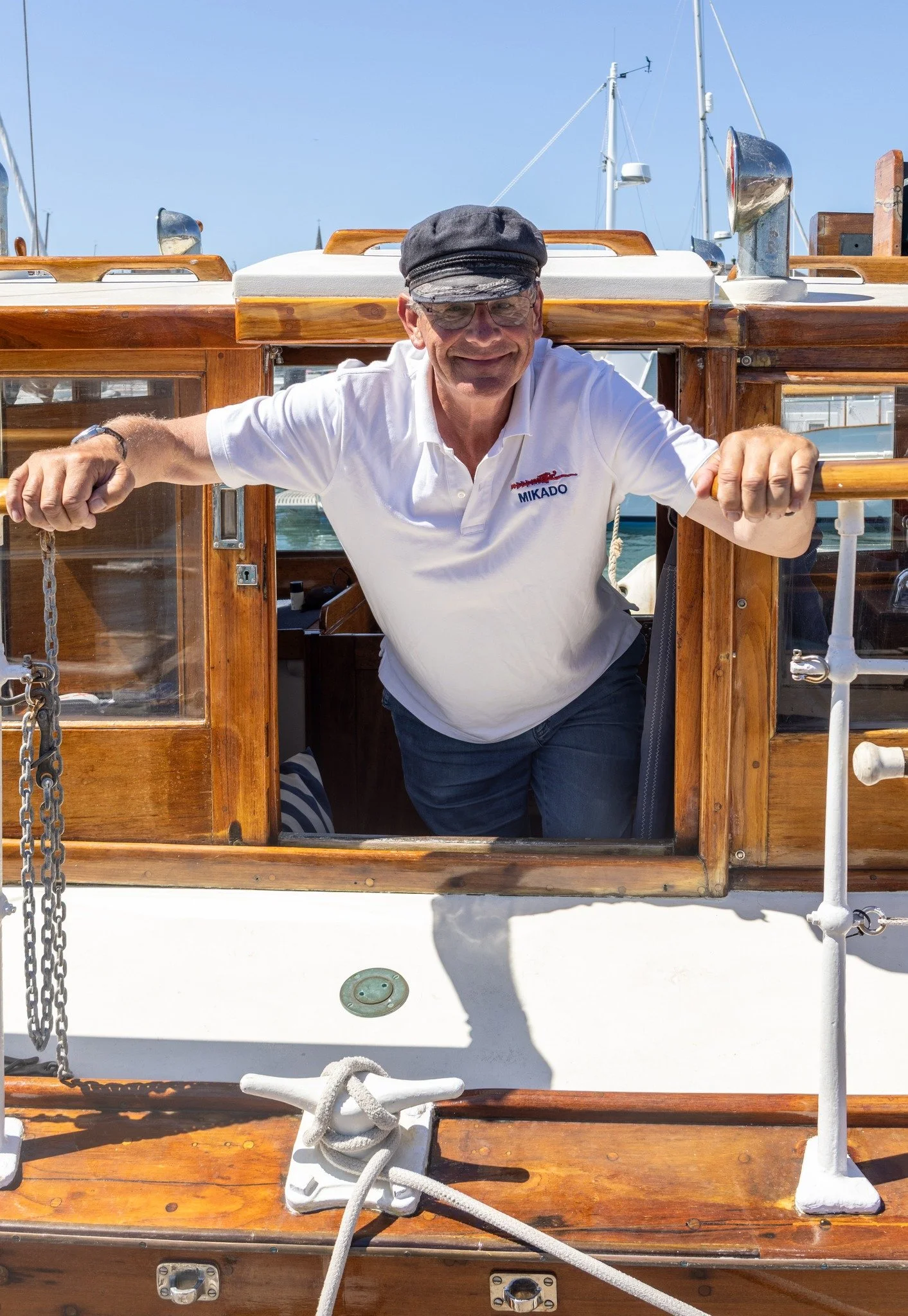 A man with glasses, wearing a black cap and white polo shirt, stands on a wooden boat, leaning through an open window with both arms out, smiling at the camera, with nautical equipment and other boats in the background.