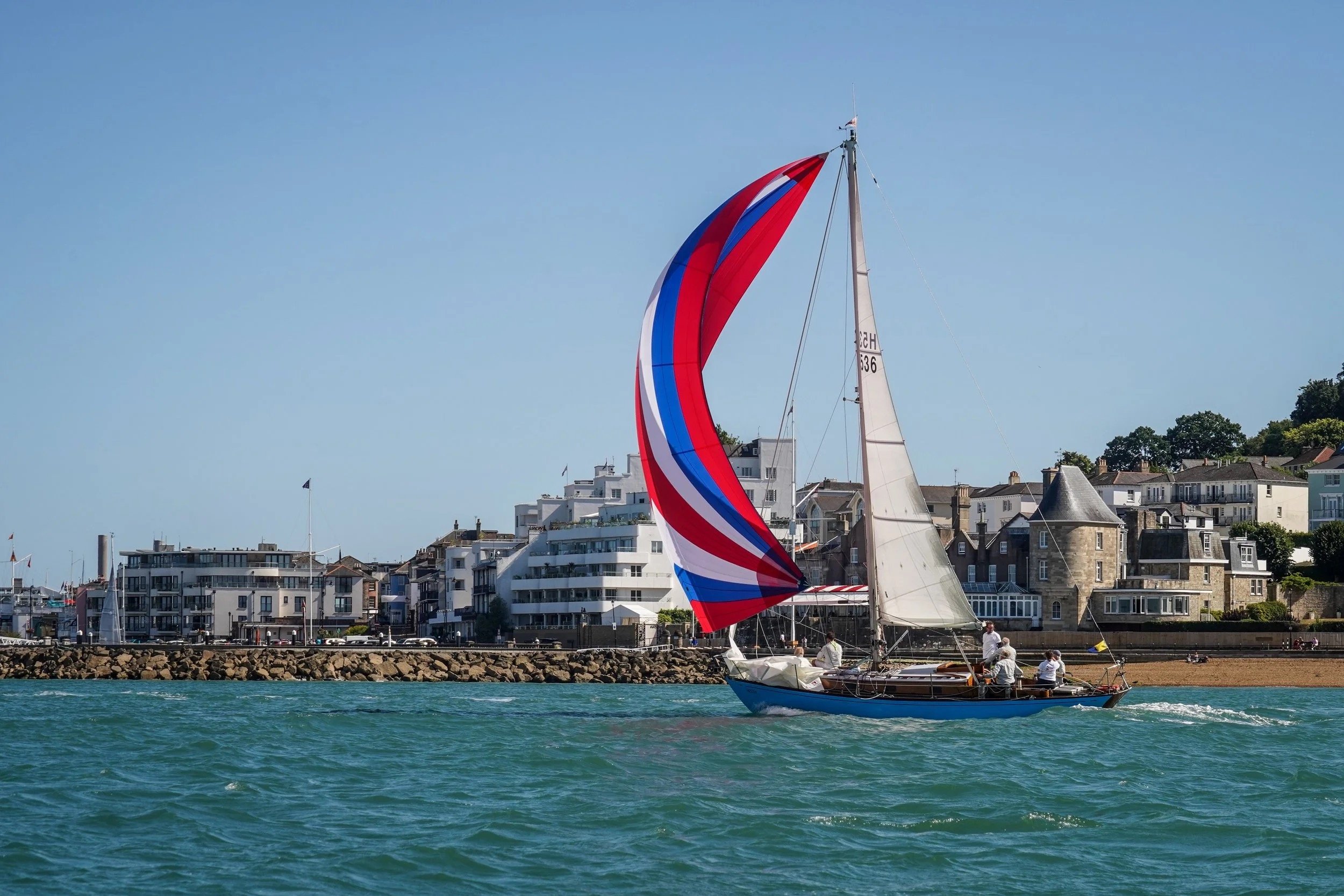 A sailboat with colorful red, blue, and white spinnaker sails on the water near a coastal town with modern white buildings and historic stone structures under a clear blue sky.