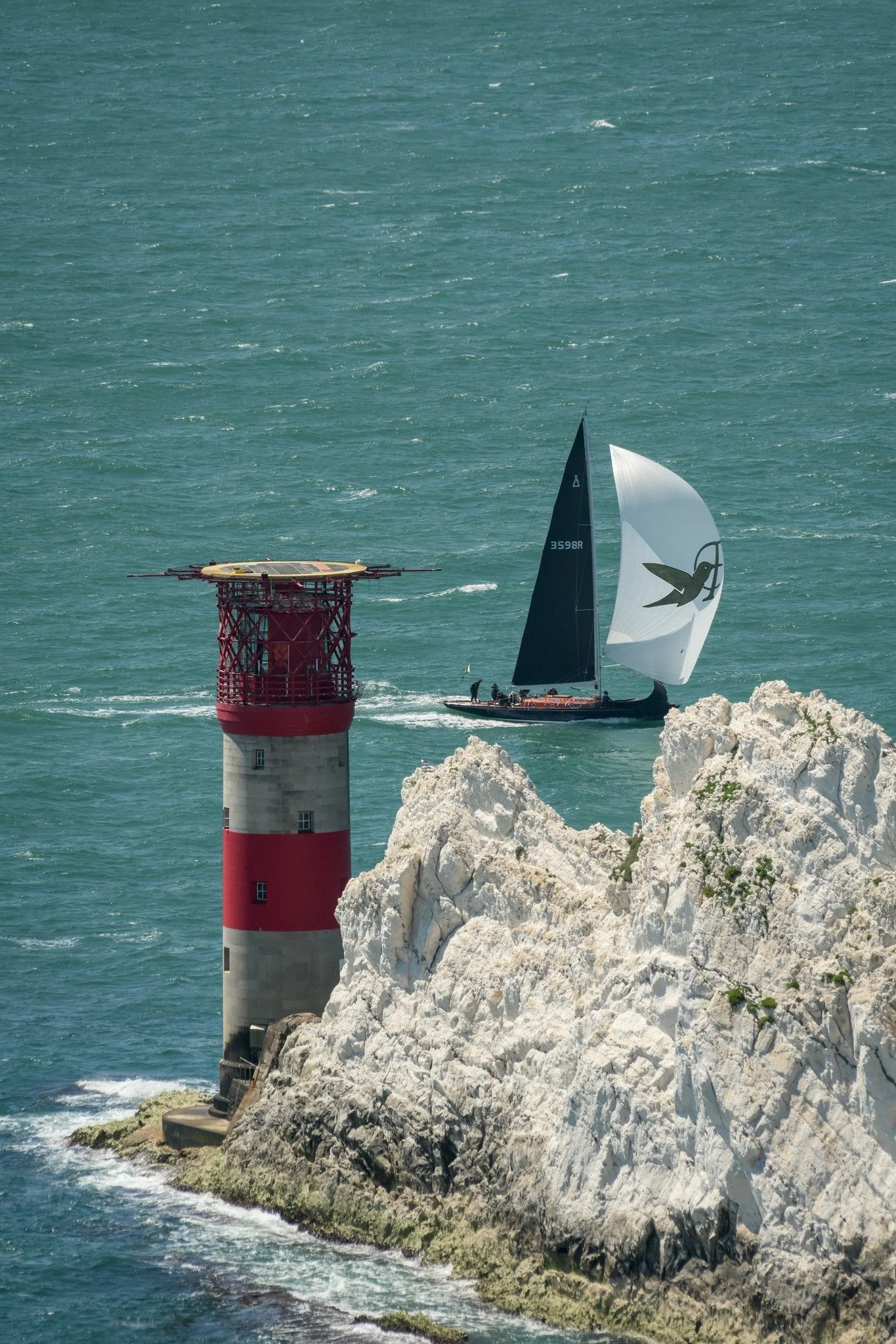 A black sailboat with a white sail featuring a bird logo sailing near a red and white striped lighthouse on rocky cliffs, with a blue ocean in the background.