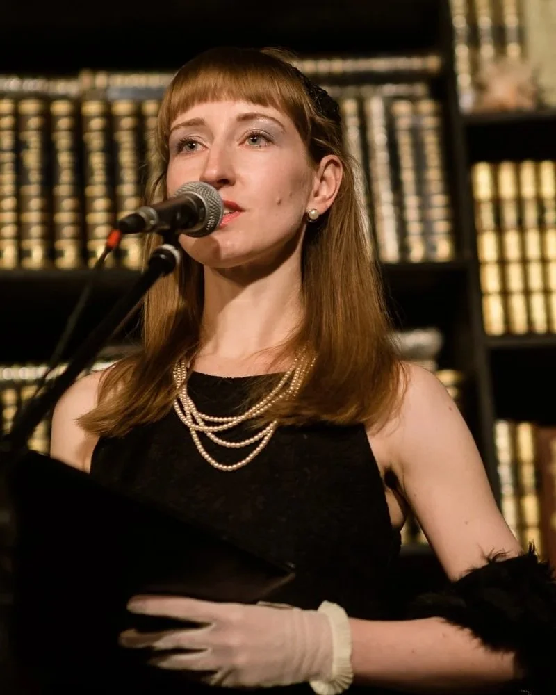 A woman with long brown hair singing into a microphone in front of a bookshelf.