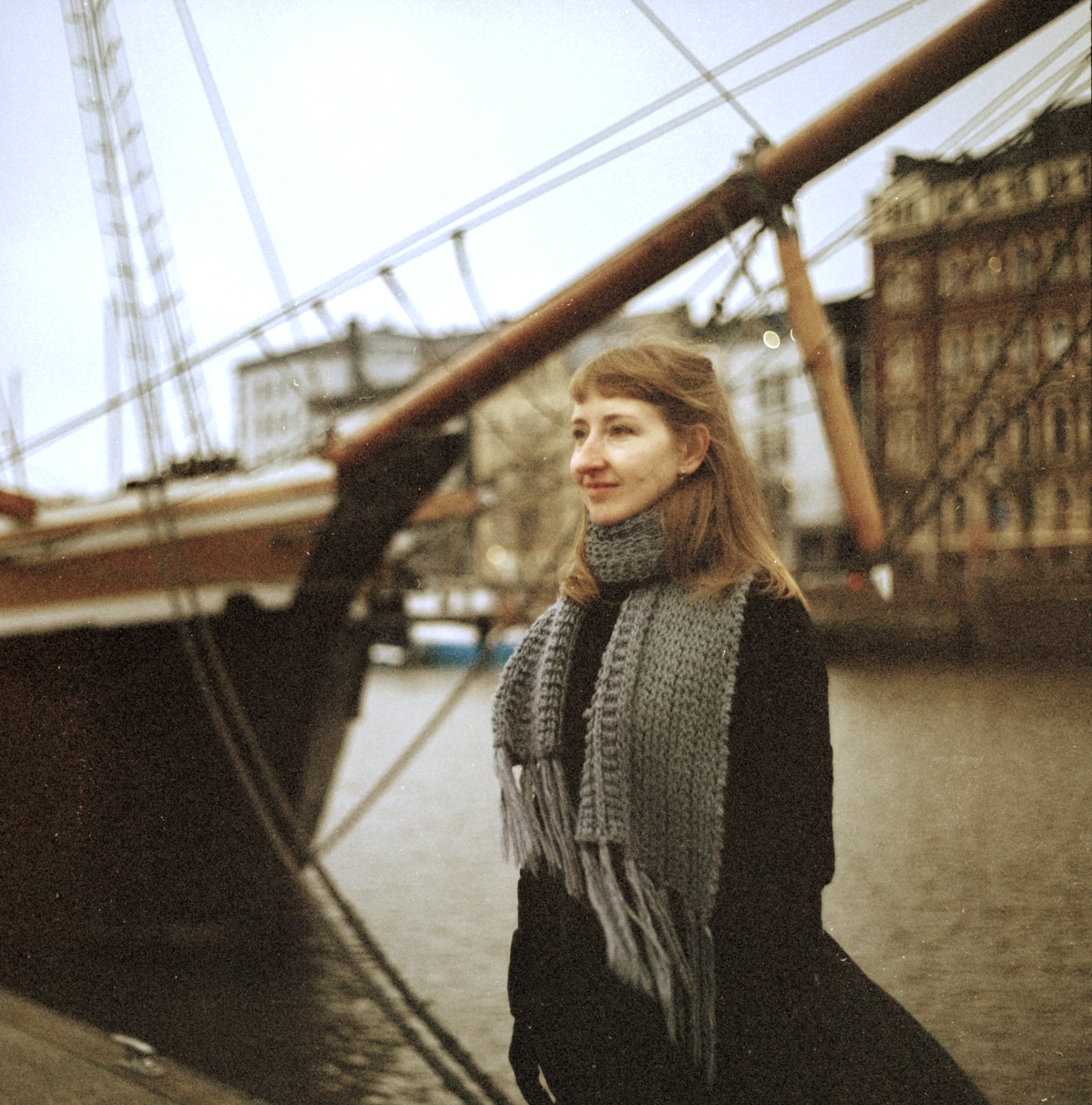 A woman with brown hair wearing a black coat and gray scarf standing near a boat docked on a river. In the background, there are historic buildings and a large Ferris wheel.