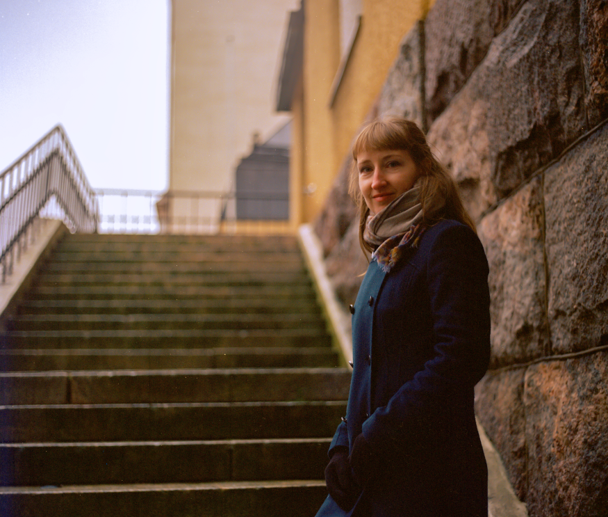 A young woman with light brown hair standing on a stone staircase outdoors, wearing a dark coat and a light scarf, smiling at the camera.
