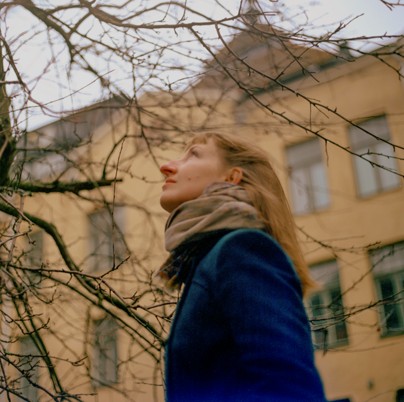 A woman with red hair wearing a blue coat and a beige scarf looking up at leafless tree branches in front of a yellow building.
