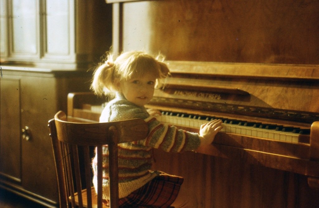 A young girl with blonde hair in pigtails sitting at an upright wooden piano, looking over her shoulder at the camera.
