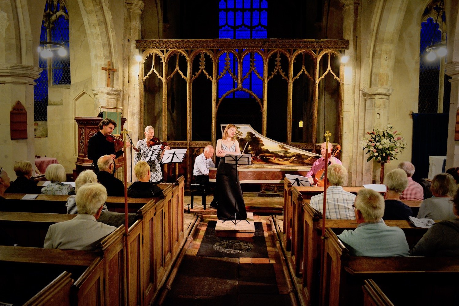 A small musical concert taking place inside a church with an audience of mainly elderly people. Musicians include a pianist, a violinist, a cellist, and a singer performing in front of a large, ornate wooden backdrop.