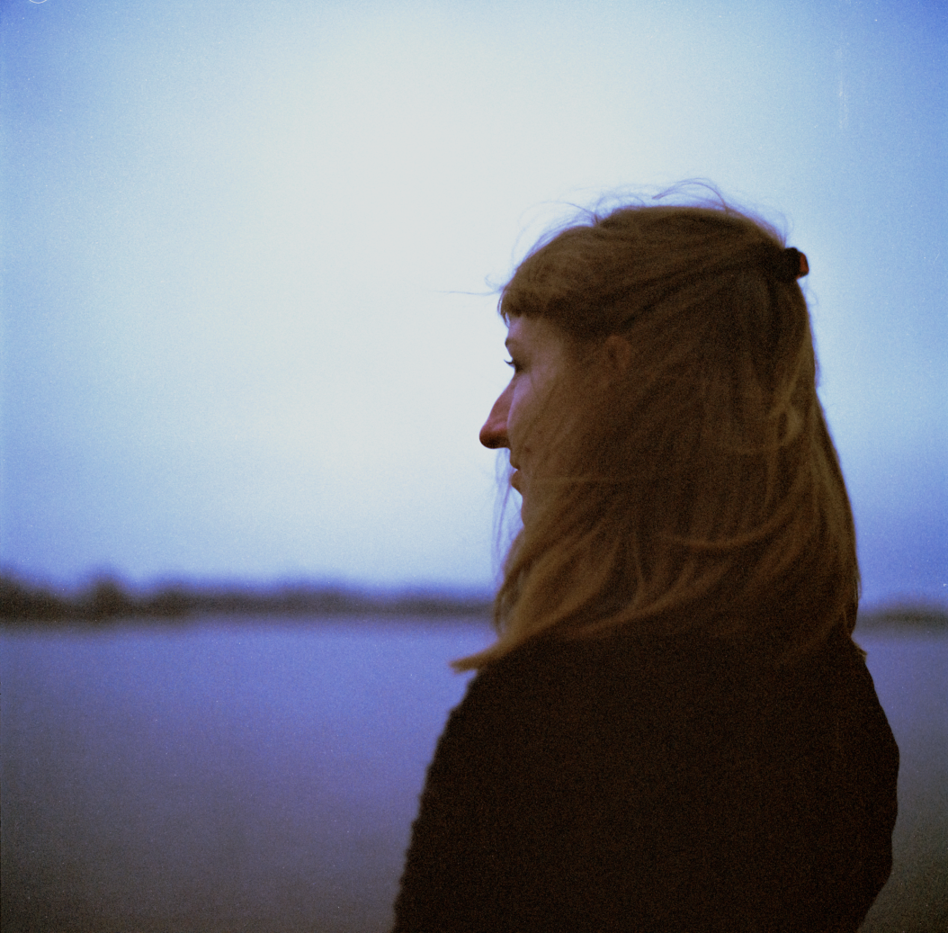 Profile of a woman with reddish-brown hair, standing near a body of water during dusk or dawn, with a clear sky in the background.