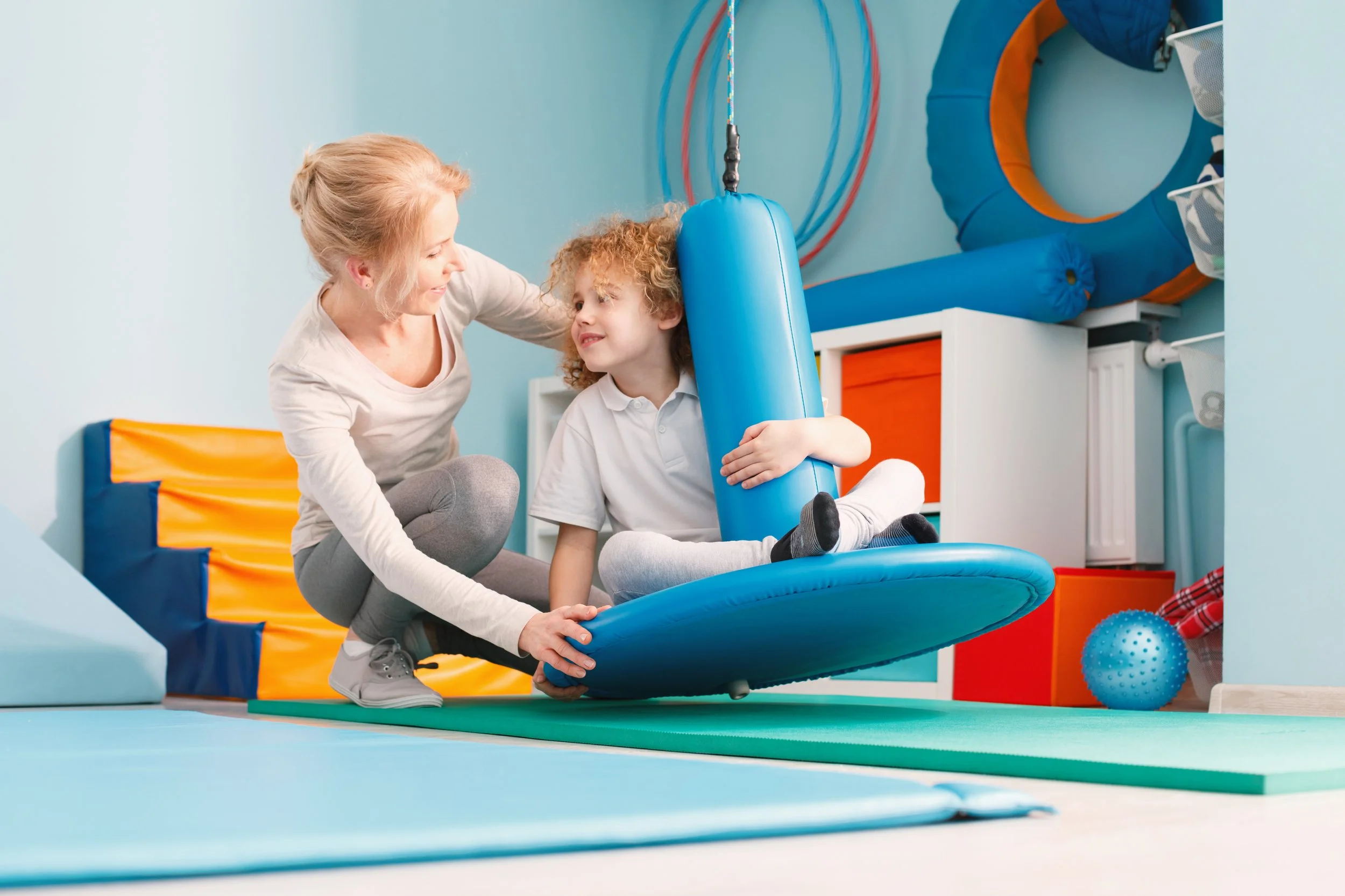 A woman helping a young boy with curly hair on a blue circular indoor playground equipment in a playroom.