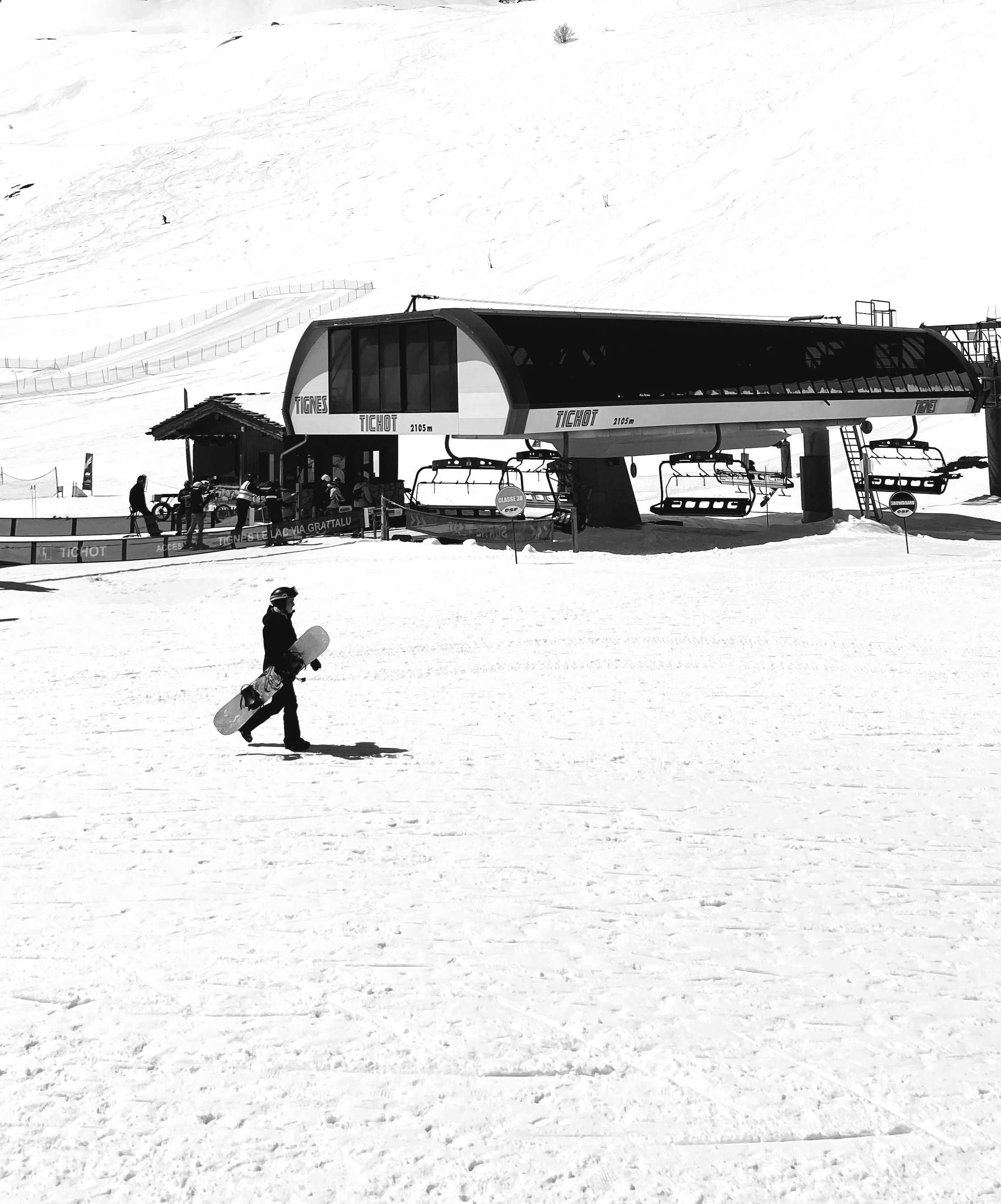 Une station de ski avec un télésiège en arrière-plan, un skieur avec une planche à neige marche sur la neige en premier plan.