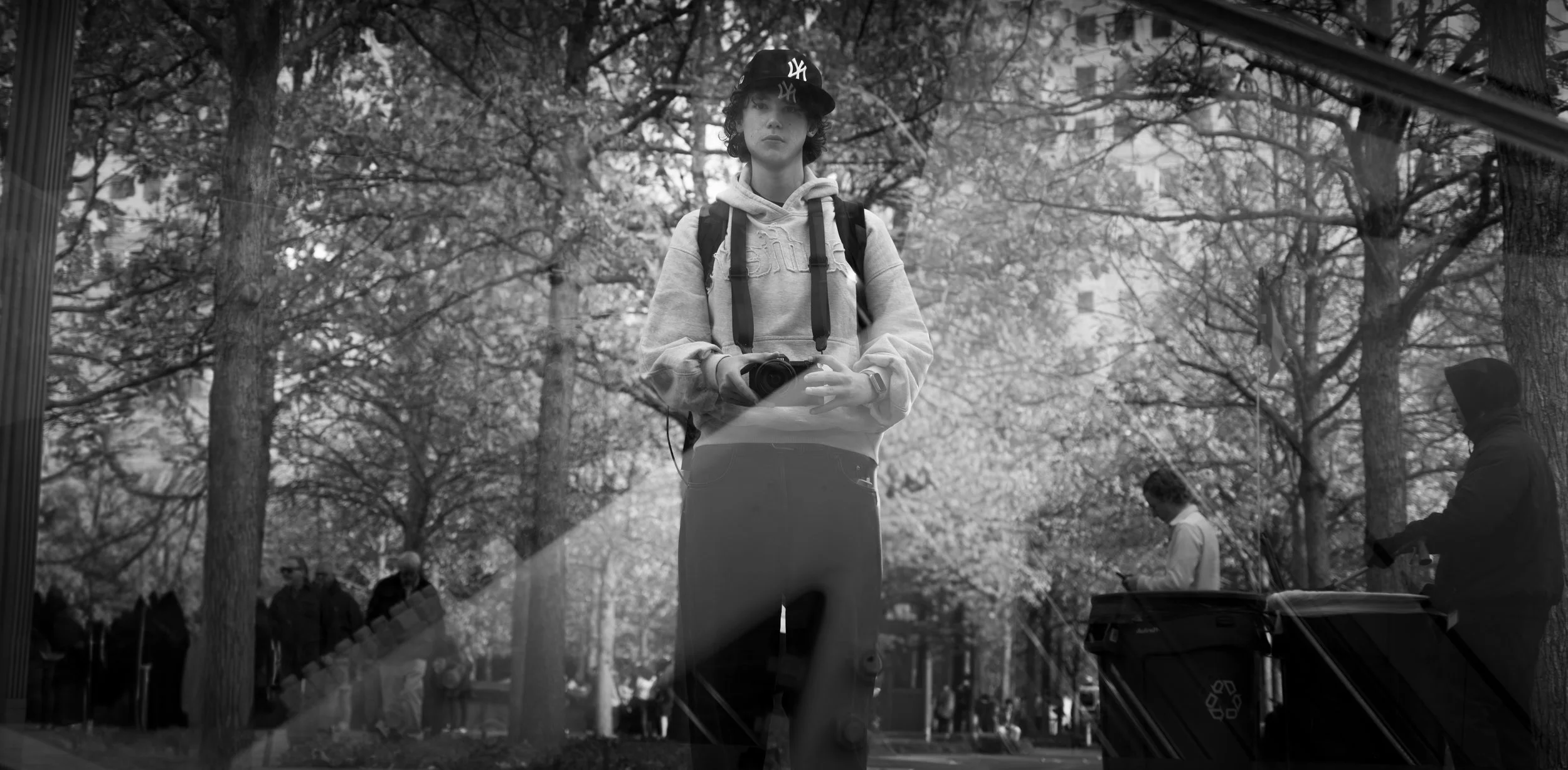 Une jeune femme rafraîchie portant un hoodie, un sac à dos et un casque de baseball, tenant un appareil photo, photographiant dans un parc avec des arbres en arrière-plan, vue à travers une vitrine reflective.