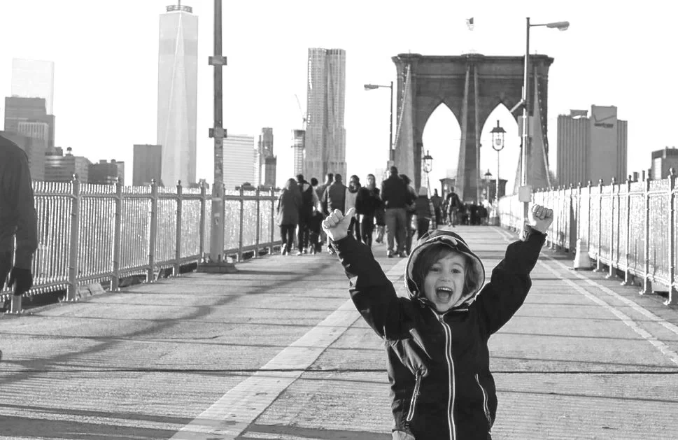 Un enfant joyeux avec les bras levés sur le pont de Brooklyn à New York, avec la skyline de Manhattan en arrière-plan.
