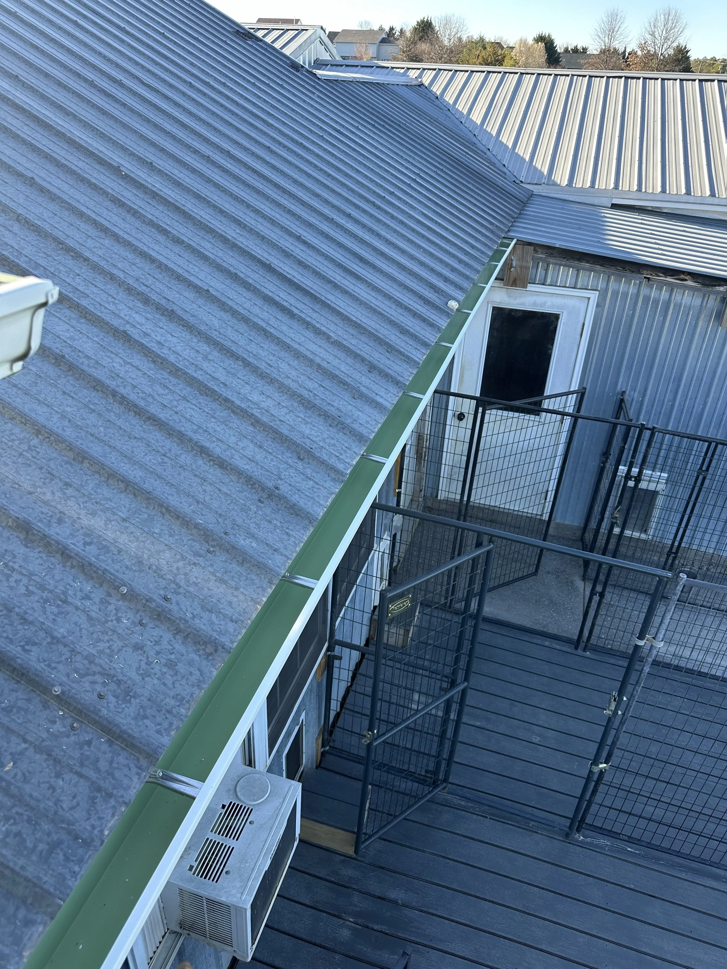 A rooftop with metal roofing, a balcony with black metal railing, a white door with glass, an air conditioning unit on the balcony, and neighboring houses in the background.