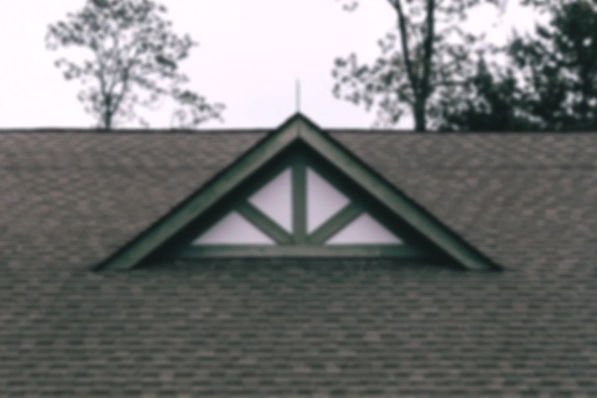Close-up of a house roof with a triangular attic vent with decorative trim, trees in the background, overcast sky.