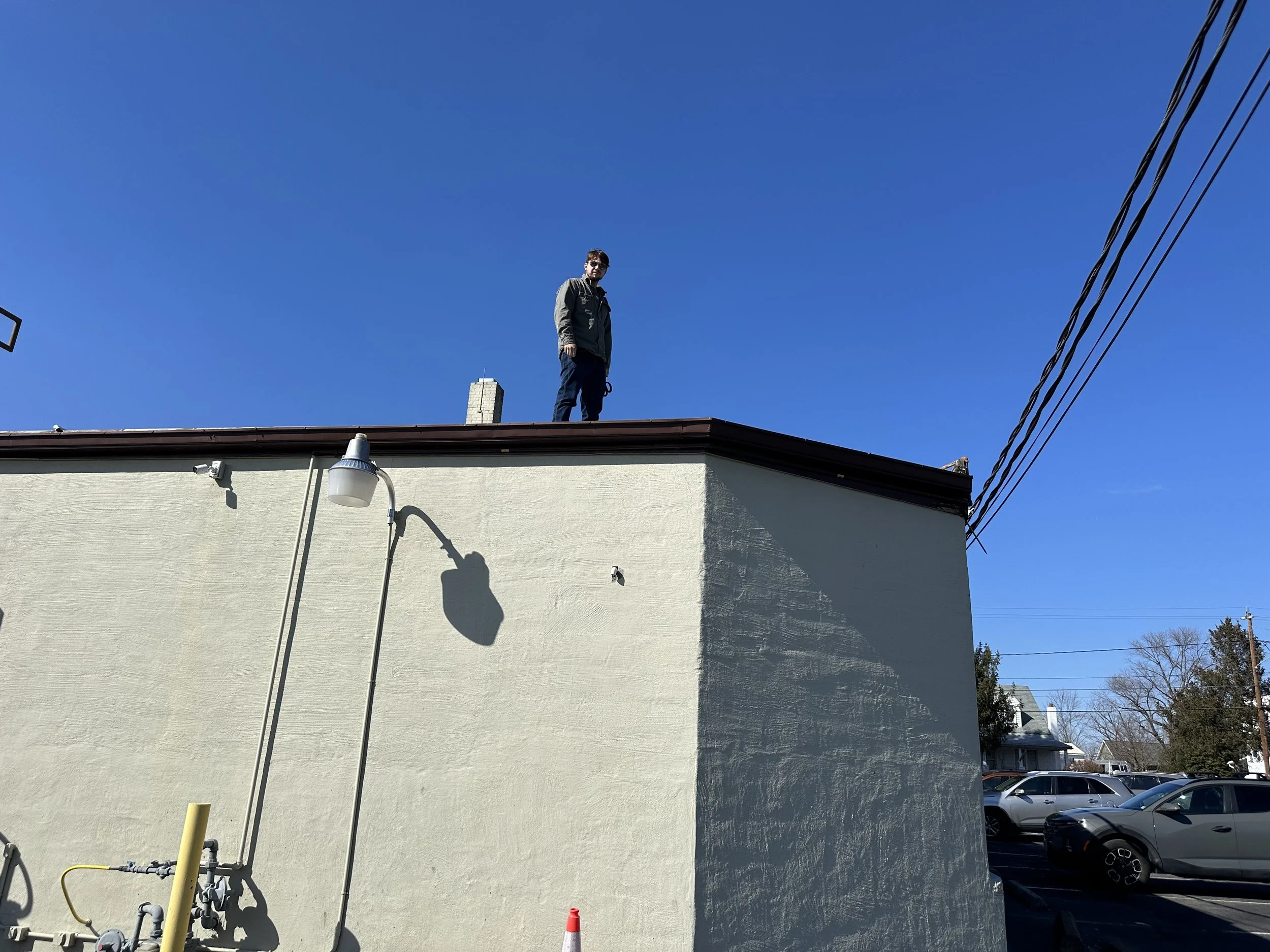 A person standing on the flat roof of a beige building, with a bright blue sky in the background and a parking lot with cars and trees nearby.