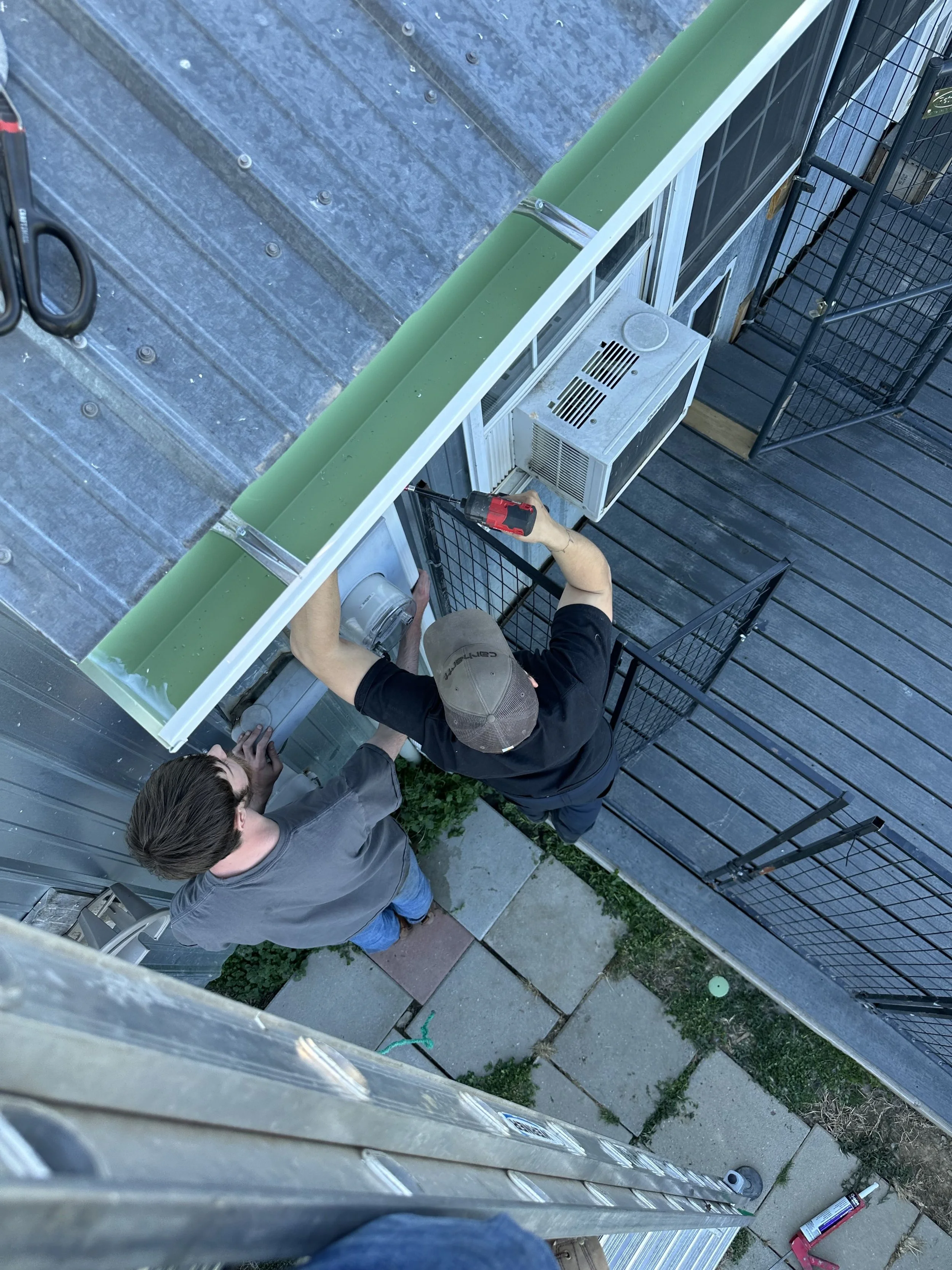 Two men working on an air conditioning unit outside a building, one of them using a power drill.
