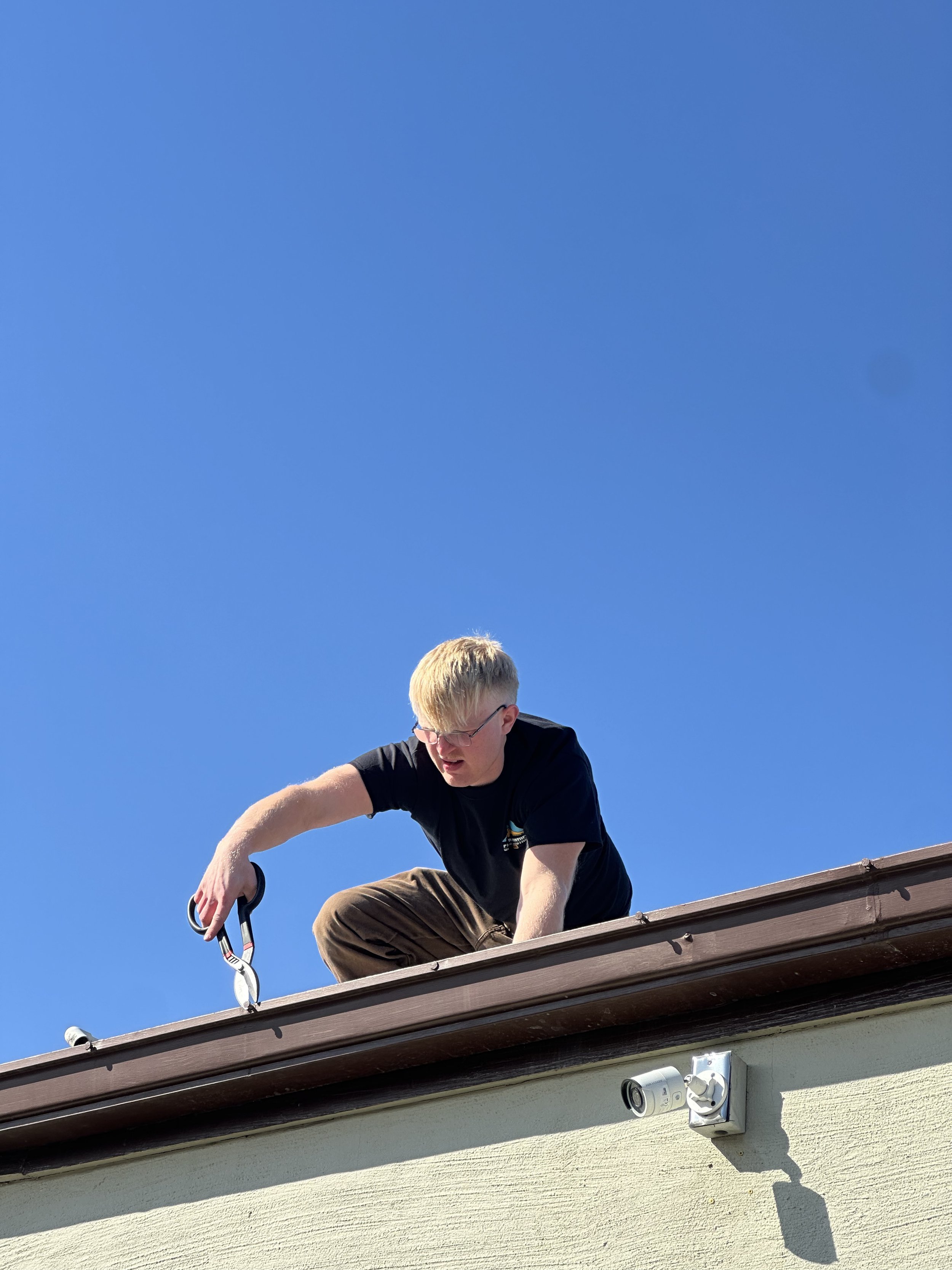 A man with glasses wearing a black T-shirt and brown pants kneels on a roof, using pruning shears, with a clear blue sky in the background.