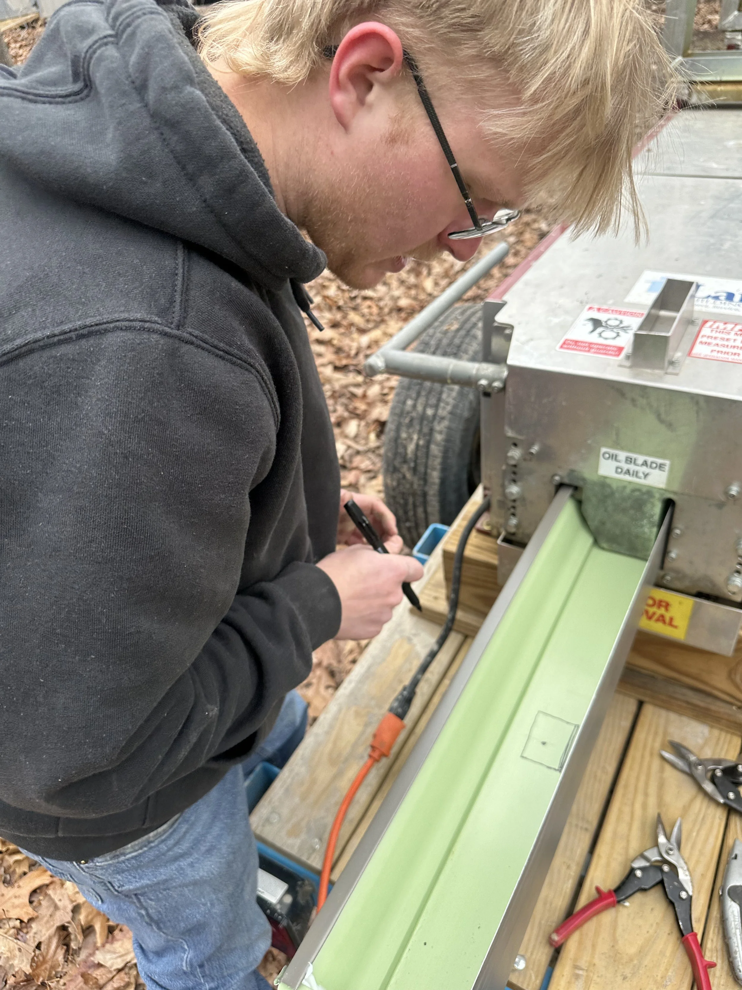 A young man with blonde hair, glasses, and a black hoodie looking down at a green channel on a metal machine outdoors surrounded by tools