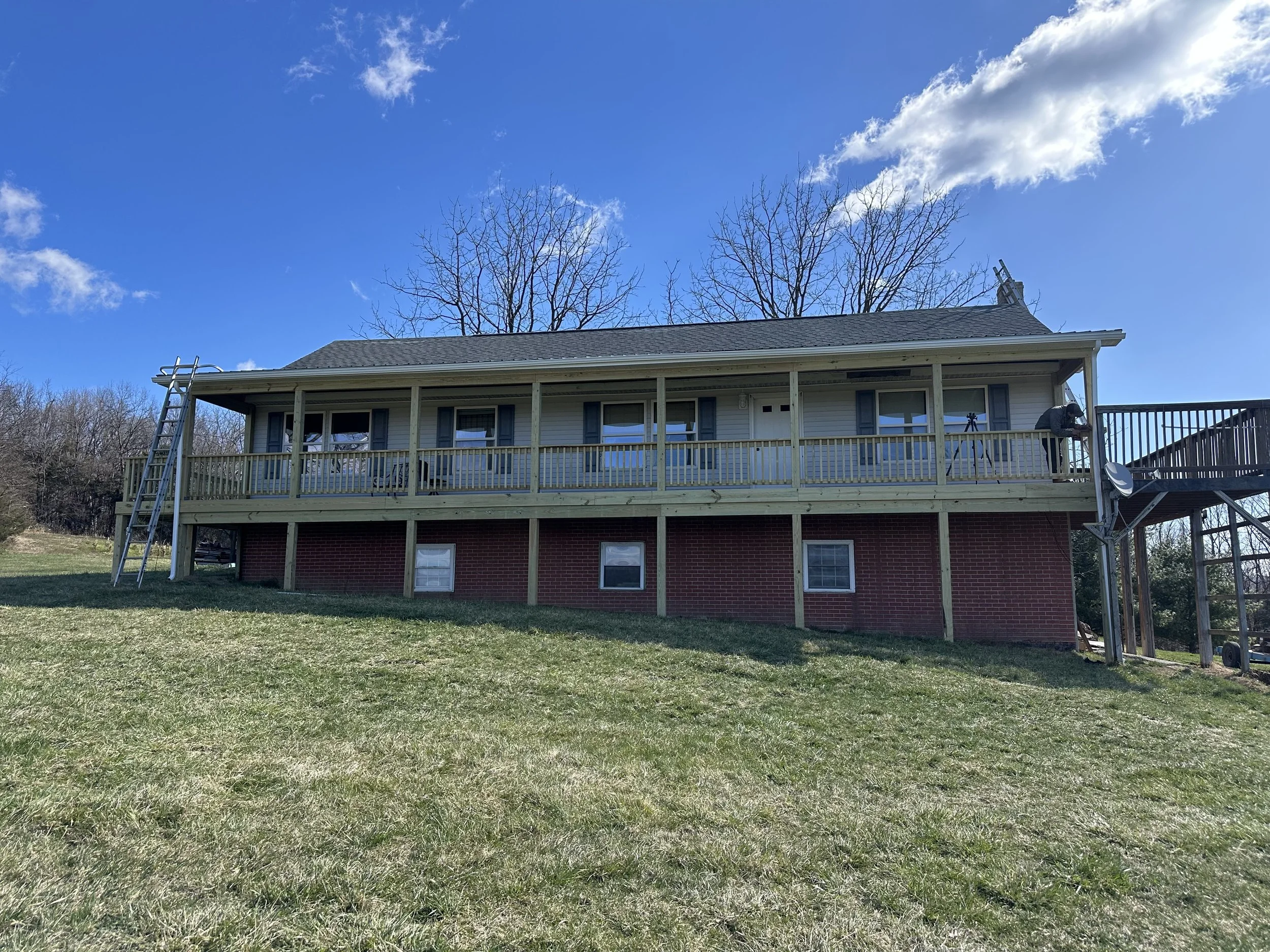 A two-story house with a red brick lower level and a light-colored upper level with a deck. The deck has a railing, a satellite dish, and a person working on a tripod. A ladder leans against the deck, and a staircase is on the right side. There are t