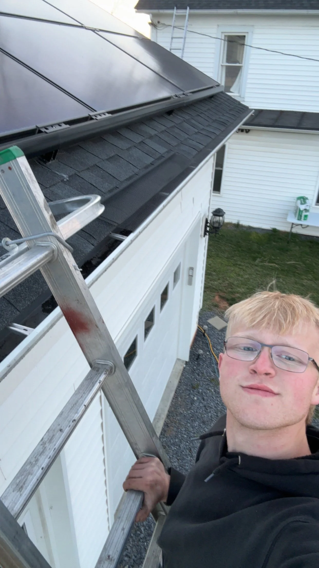 A young man with glasses taking a selfie on a ladder, working on installing solar panels on the roof of a white house.