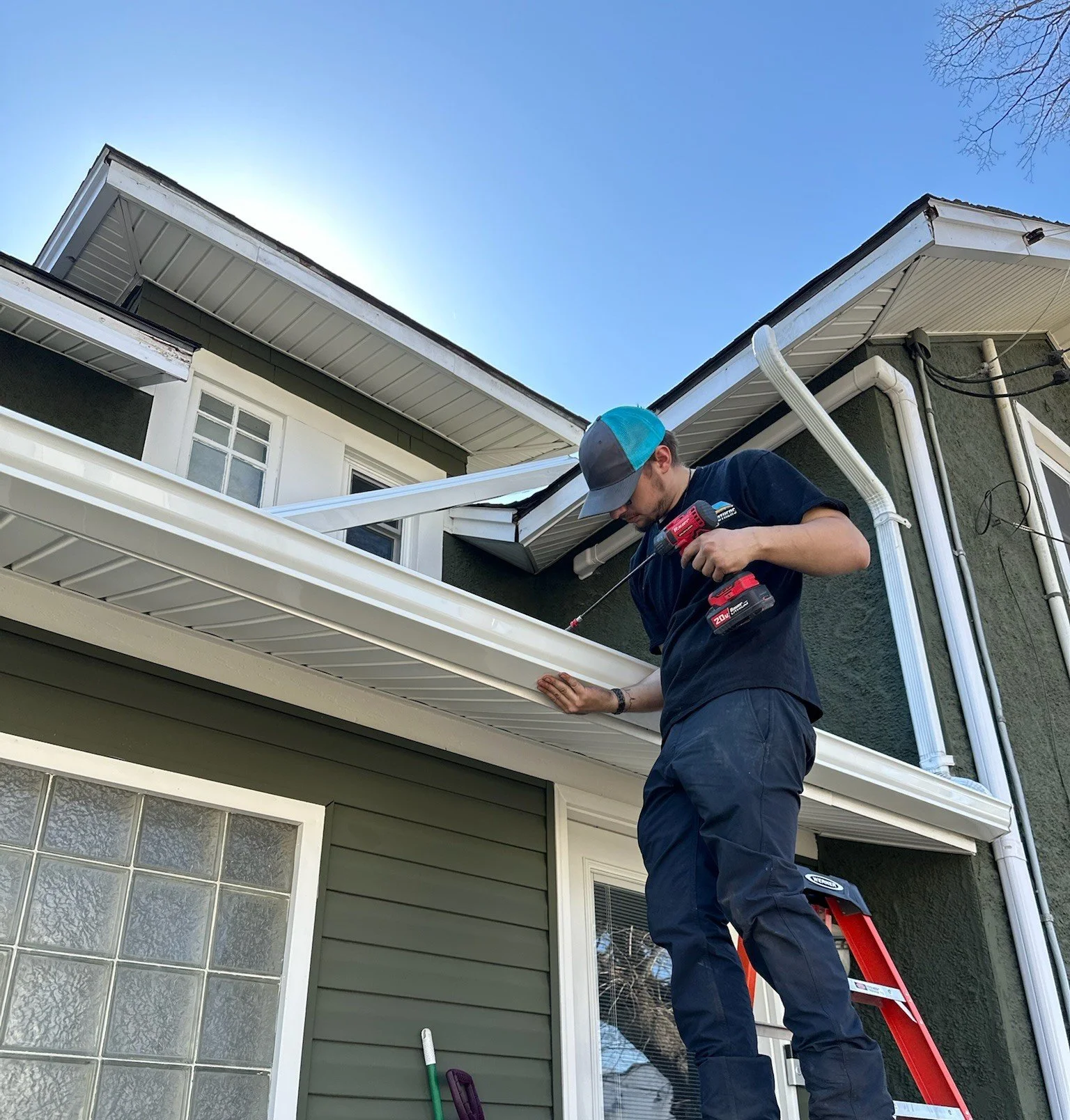 A man working on the soffit and fascia of a house, using a cordless drill, standing on a ladder, under clear blue sky.