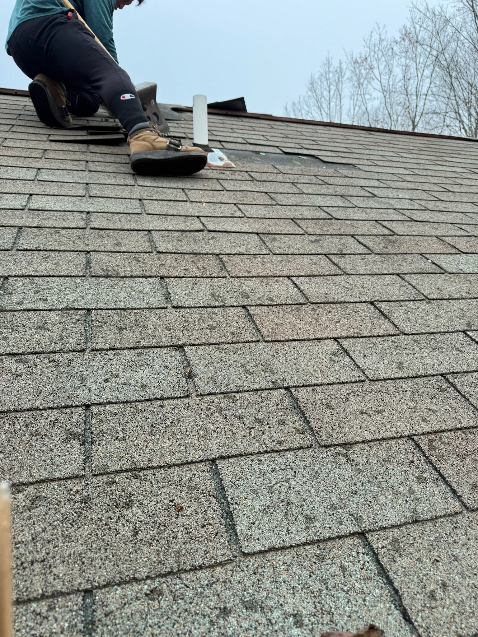 Person repairing or inspecting a shingled roof, kneeling on the roof with tools, near a white vent pipe, with trees and an overcast sky in the background.