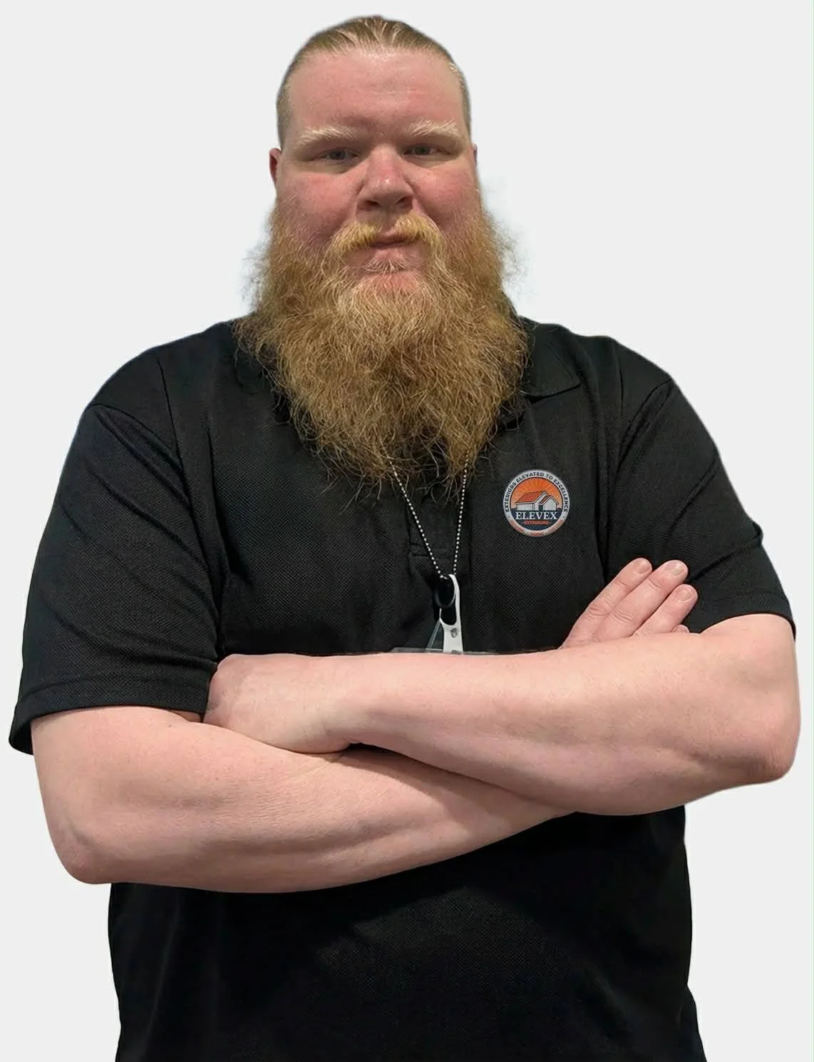 A man with a red beard and mustache, wearing a black shirt with a logo, crosses his arms and looks at the camera against a plain background.