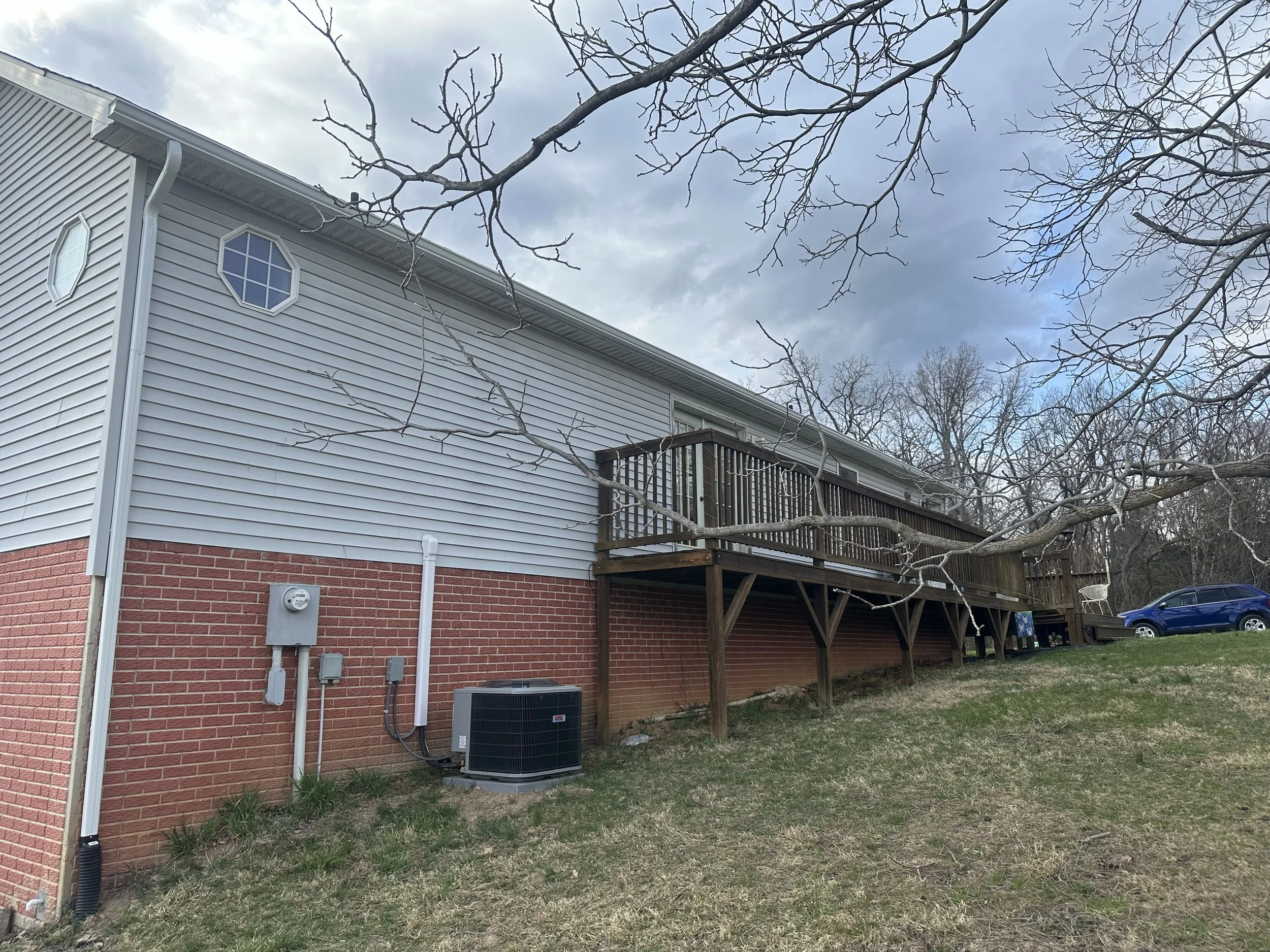 Side view of a house with a brick lower section and vinyl siding upper section, a wooden deck on stilts, and a blue car parked on a grassy area. Bare tree branches are in the foreground.