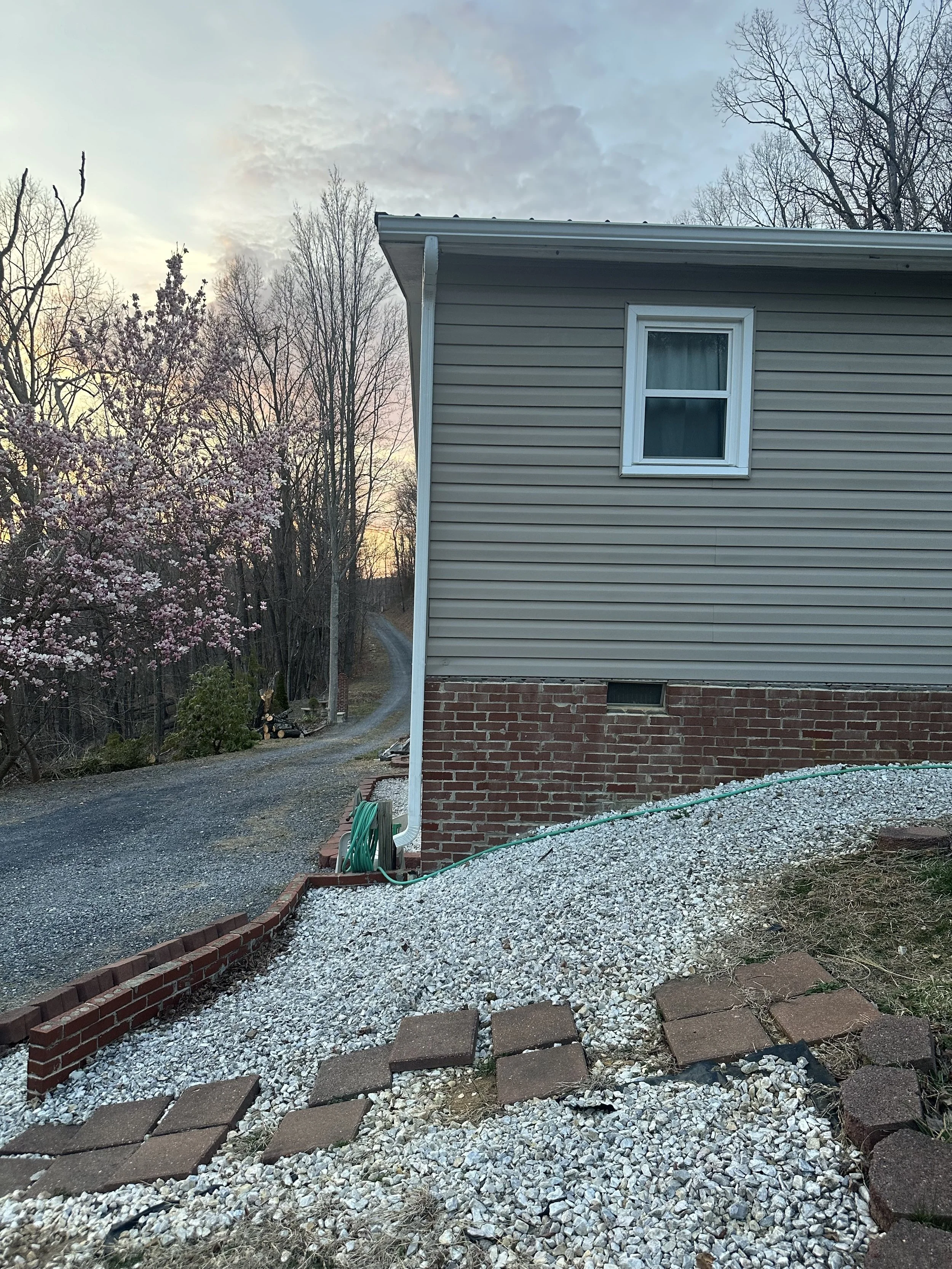 Side view of a house with beige siding and brick foundation, with a small rectangular window, a green garden hose, and a gravel pathway bordered by bricks, leading to a driveway with trees and flowering bushes in the background during sunset.