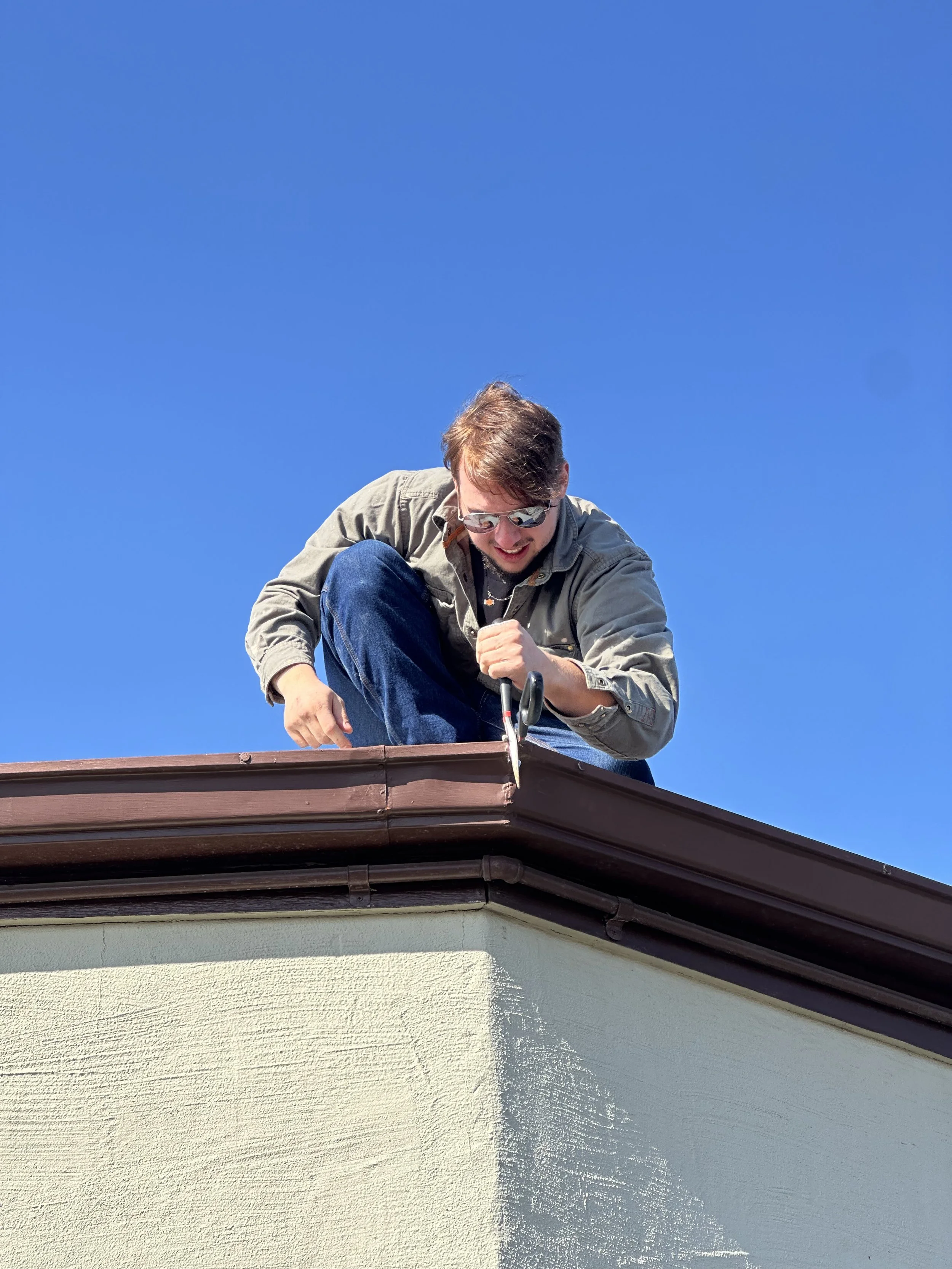 A man with sunglasses sitting on the edge of a roof, using scissors to trim or cut something with a focused expression, against a clear blue sky.