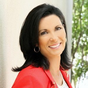 A woman with dark hair smiling, wearing a red jacket, standing outdoors with green trees in the background.
