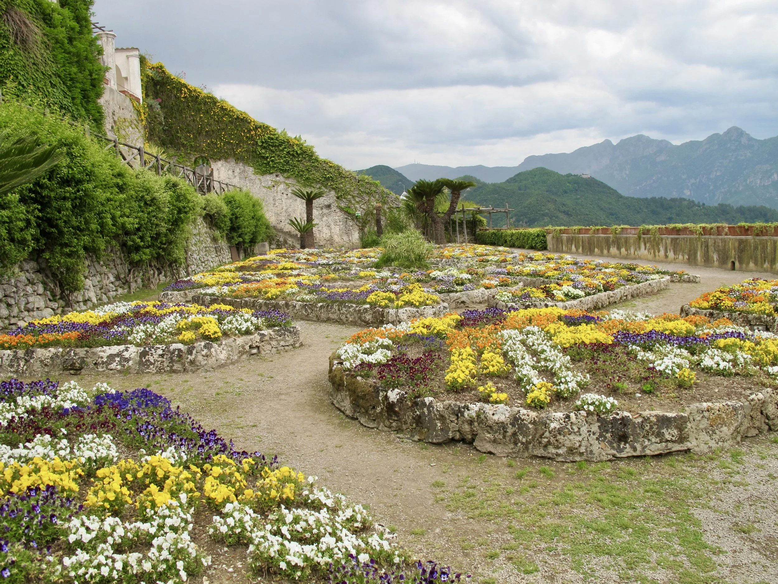 A garden with colorful flowers arranged in raised stone beds on a dirt path, surrounded by green trees and mountains in the background during cloudy weather.