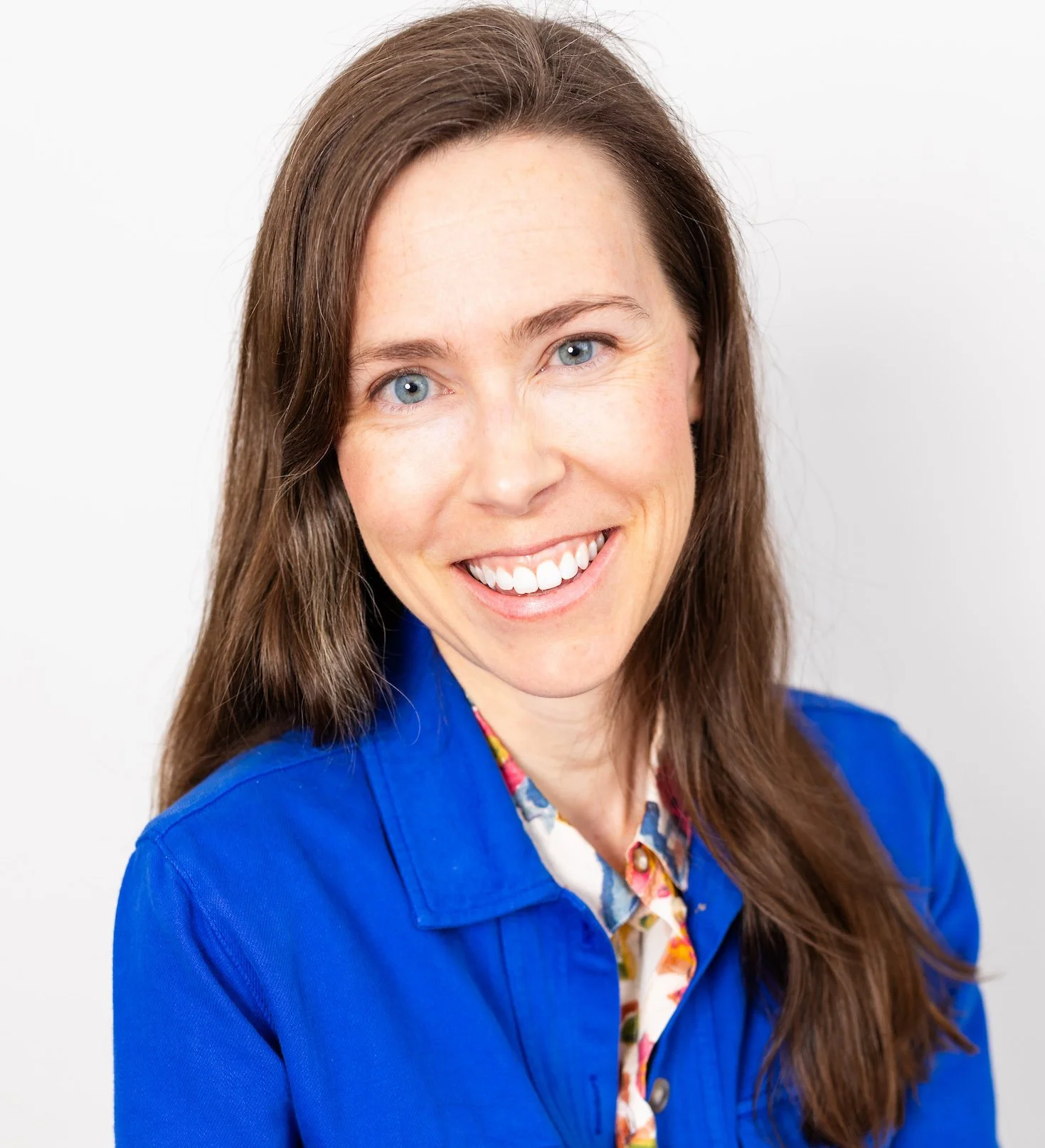 Headshot of a woman with long brown hair, blue eyes, smiling, wearing a blue jacket over a colorful floral shirt, against a white background.