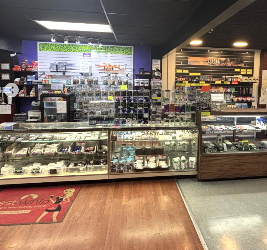 A display case filled with various petty items in a retail store, with a section of the store's wall behind it holding small products and signs, and a wooden floor with a red and black mat that says "Best Unicorn" near the entrance.