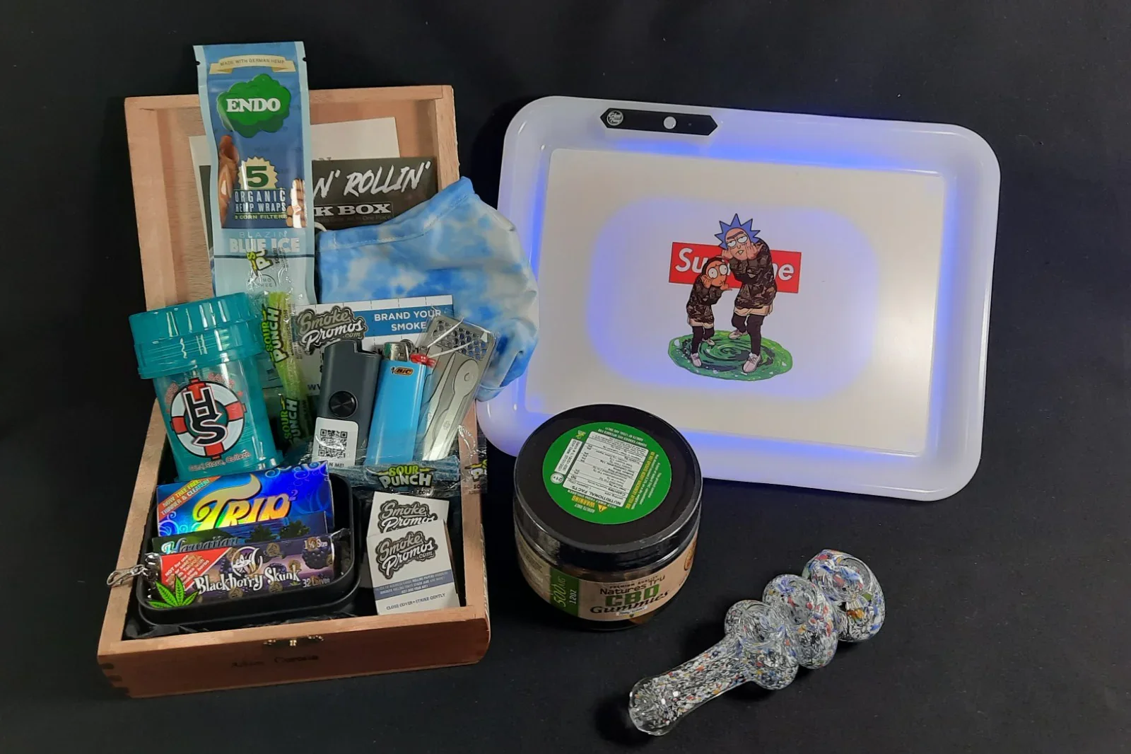 A collection of smoking accessories and cannabis products, including a jar of CBD gummies, rolling papers, lighters, a glass pipe, and pre-rolled cannabis cones, arranged on a black surface with a decorative light-up box featuring animated characters and the Supreme logo.