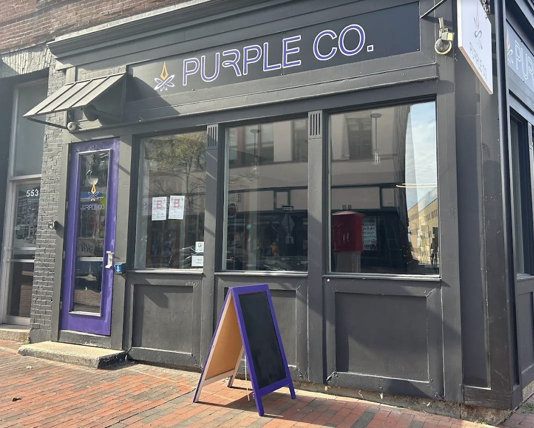 The exterior of a storefront with a sign that reads 'PURPLE CO.' The storefront has black walls, large windows, and a purple door with a small awning. There is a purple and black sandwich board sign placed outside on the red brick sidewalk.