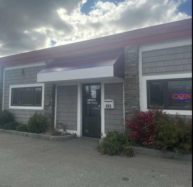 Exterior view of a medical office building with a black door, a white mailbox labeled '431,' a neon 'Open' sign in the window, and a sign on the door that says 'Medical 929-2222.' The building has gray shingles and stone accents, with bushes in front.