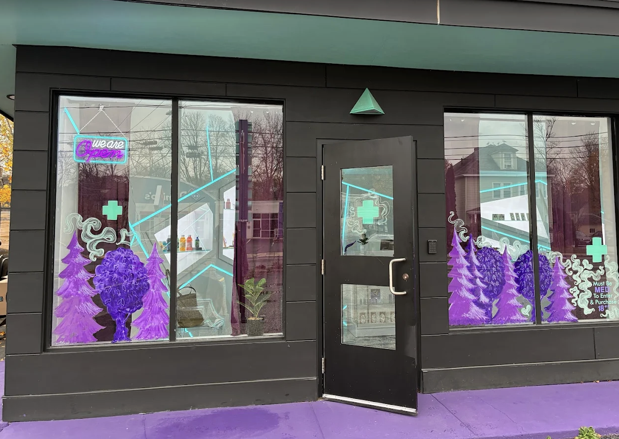 Facade of a black storefront with large glass windows decorated with purple trees, a large purple flower, a plus sign, and neon signs including one that says 'We are open.' Inside, there are shelves with bottles, a plant, and neon blue lines. The door is slightly open with a purple floor outside.