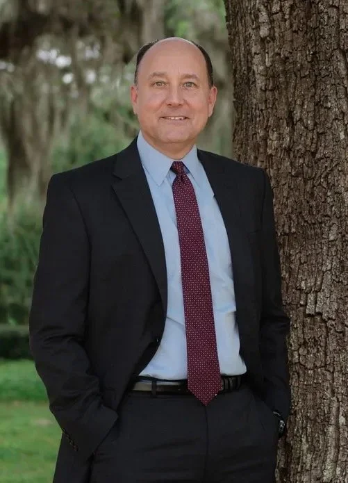 A smiling man in a dark suit and red tie standing outdoors next to a tree.