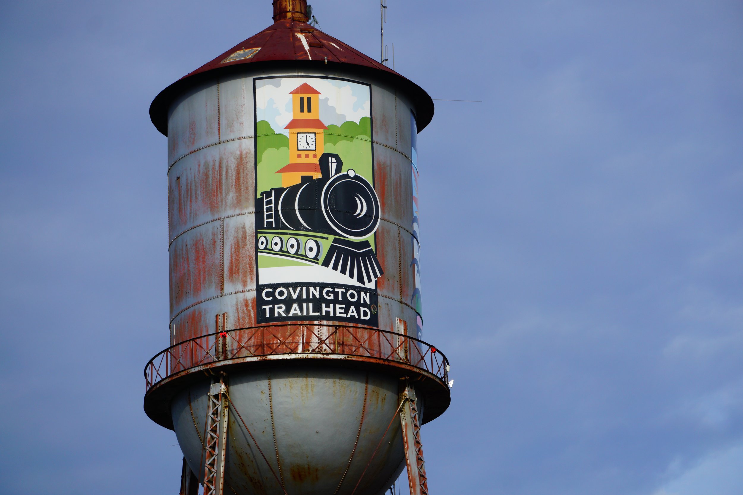 A rusty water tower with a colorful sign for Covington Trailhead featuring a black steam locomotive, a clock tower, and green trees against a blue sky.