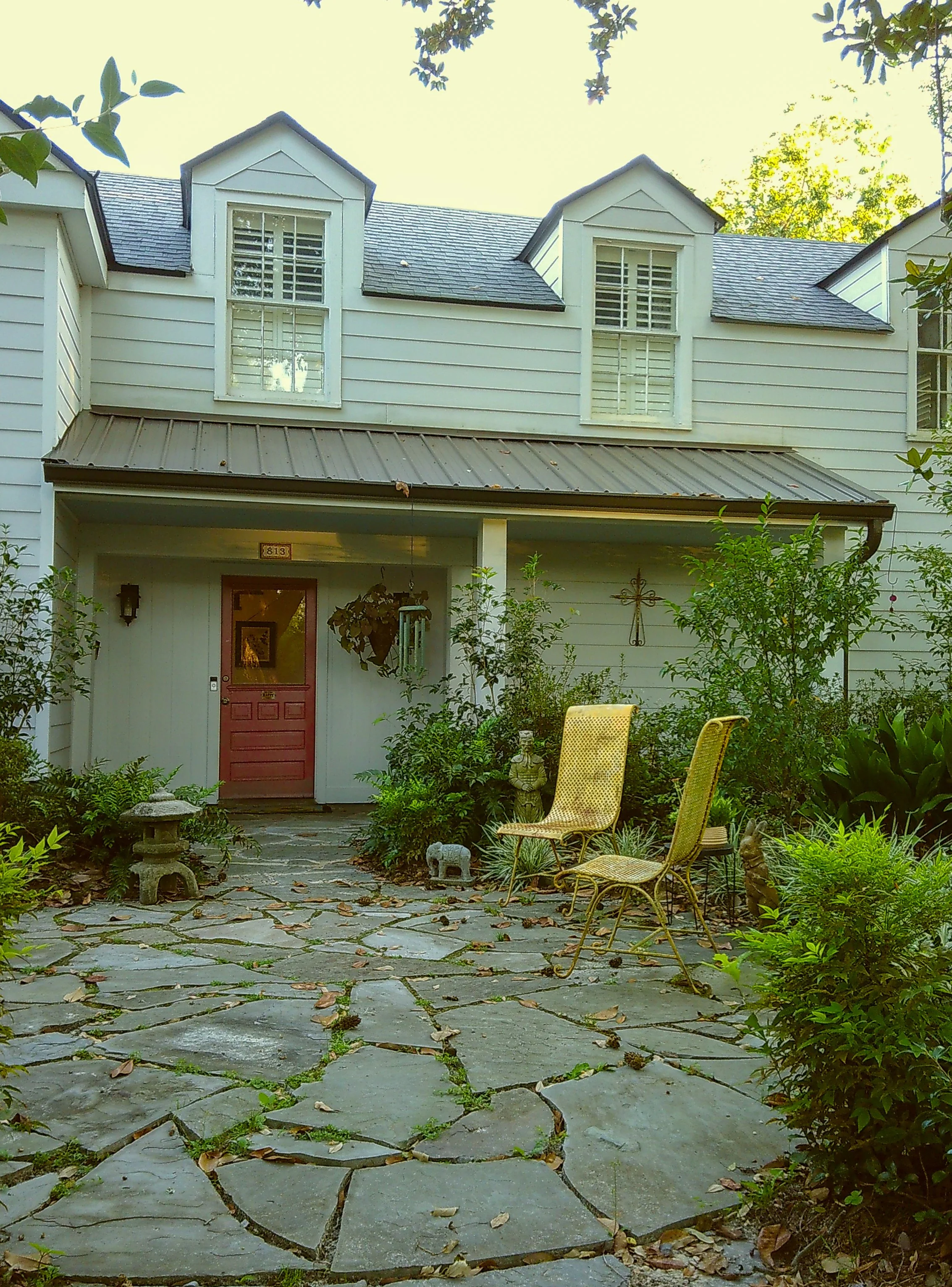 Front porch of a white house with a pink door, surrounded by greenery, two yellow chairs, garden decorations, and a stone pathway.
